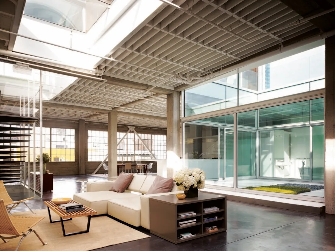 An industrial loft living room with an exposed concrete waffle ceiling, a skylight, a cream sectional sofa, and a glass-walled atrium.