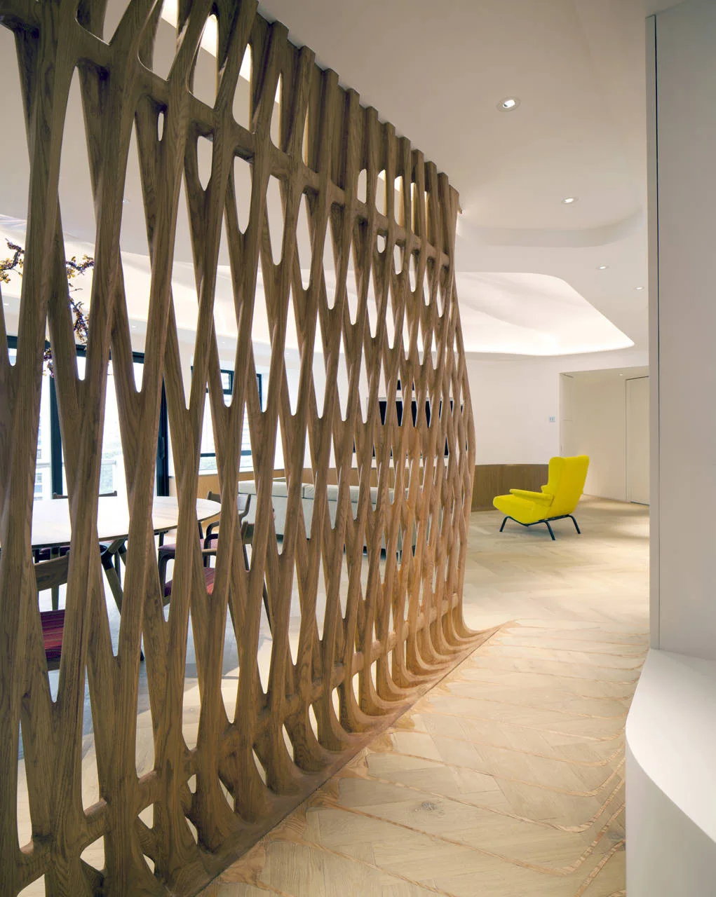 A modern open interior featuring a tall sculptural wooden lattice room divider on light herringbone floors next to a yellow accent chair.