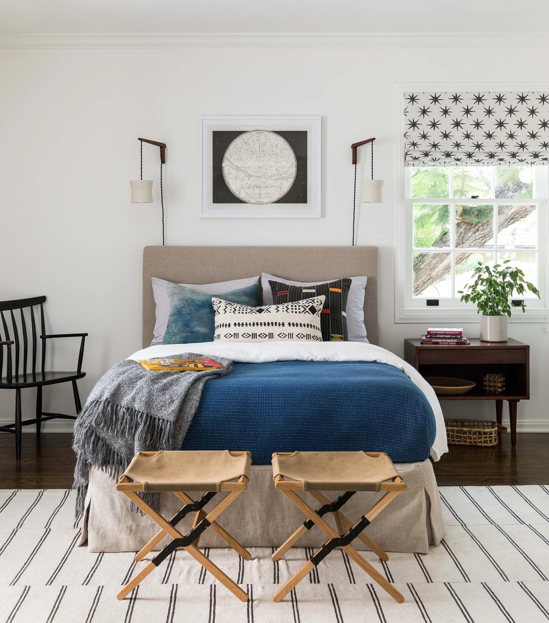 A transitional bedroom features an upholstered bed with indigo bedding, a wooden nightstand, a patterned roman shade, and two wooden folding stools.
