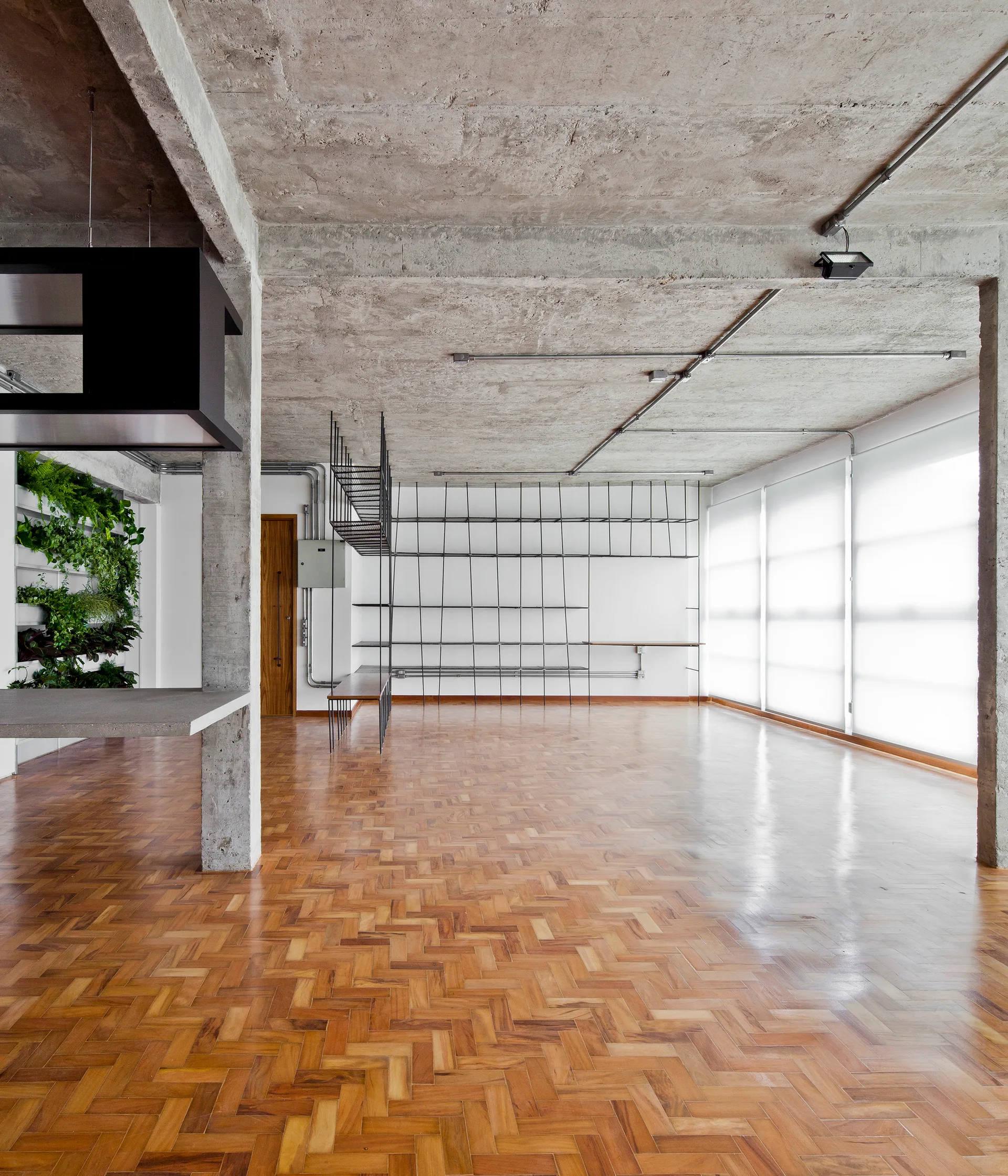 An empty loft interior featuring glossy herringbone wood floors, an exposed raw concrete ceiling and columns, and a black steel grid shelving system.