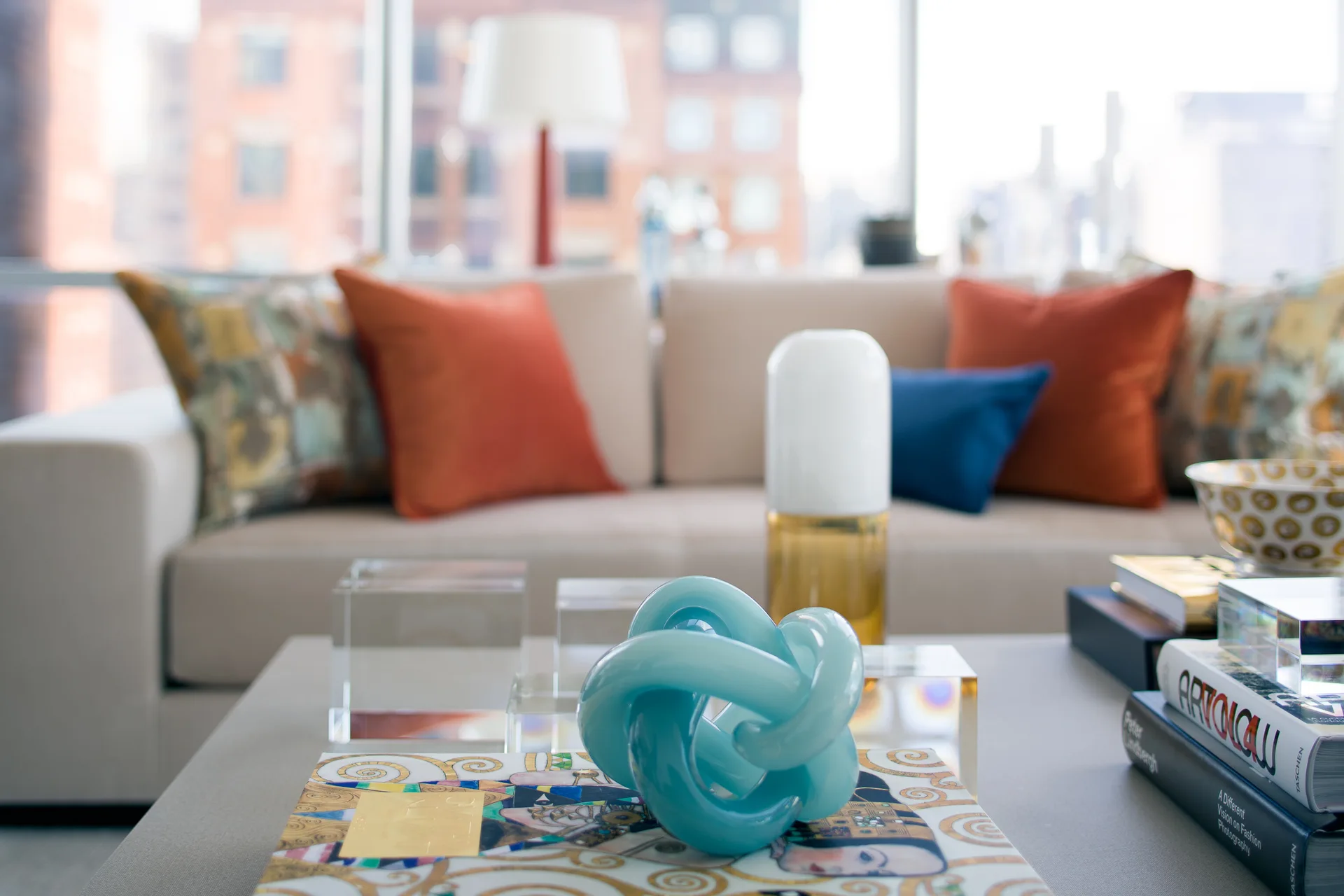 A light blue knotted glass sculpture rests on an art book atop a coffee table, in front of a beige sofa with orange and blue throw pillows.