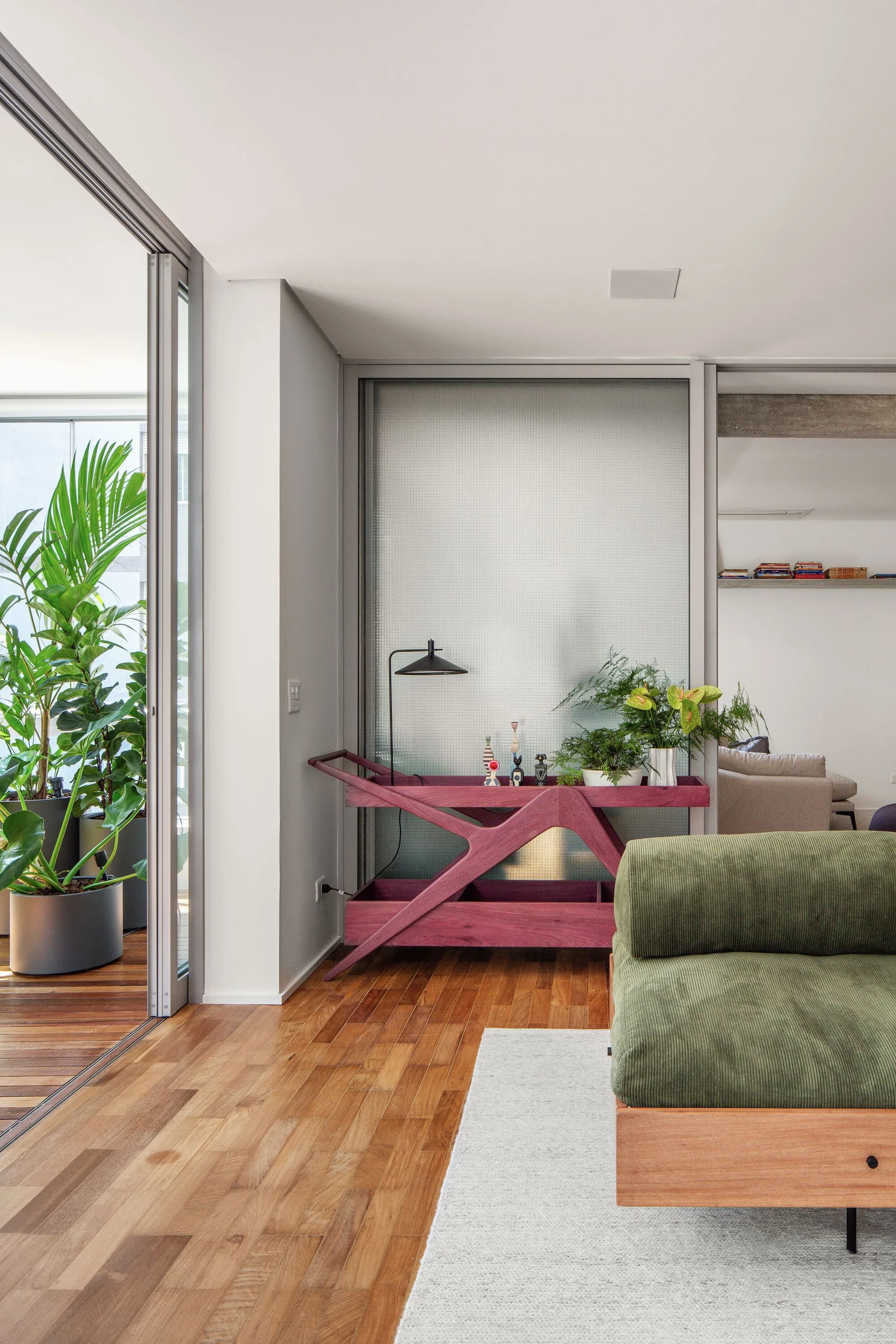 A contemporary living room features a green corduroy sofa, a plum-purple wooden console table against a textured glass wall, and wood floors.