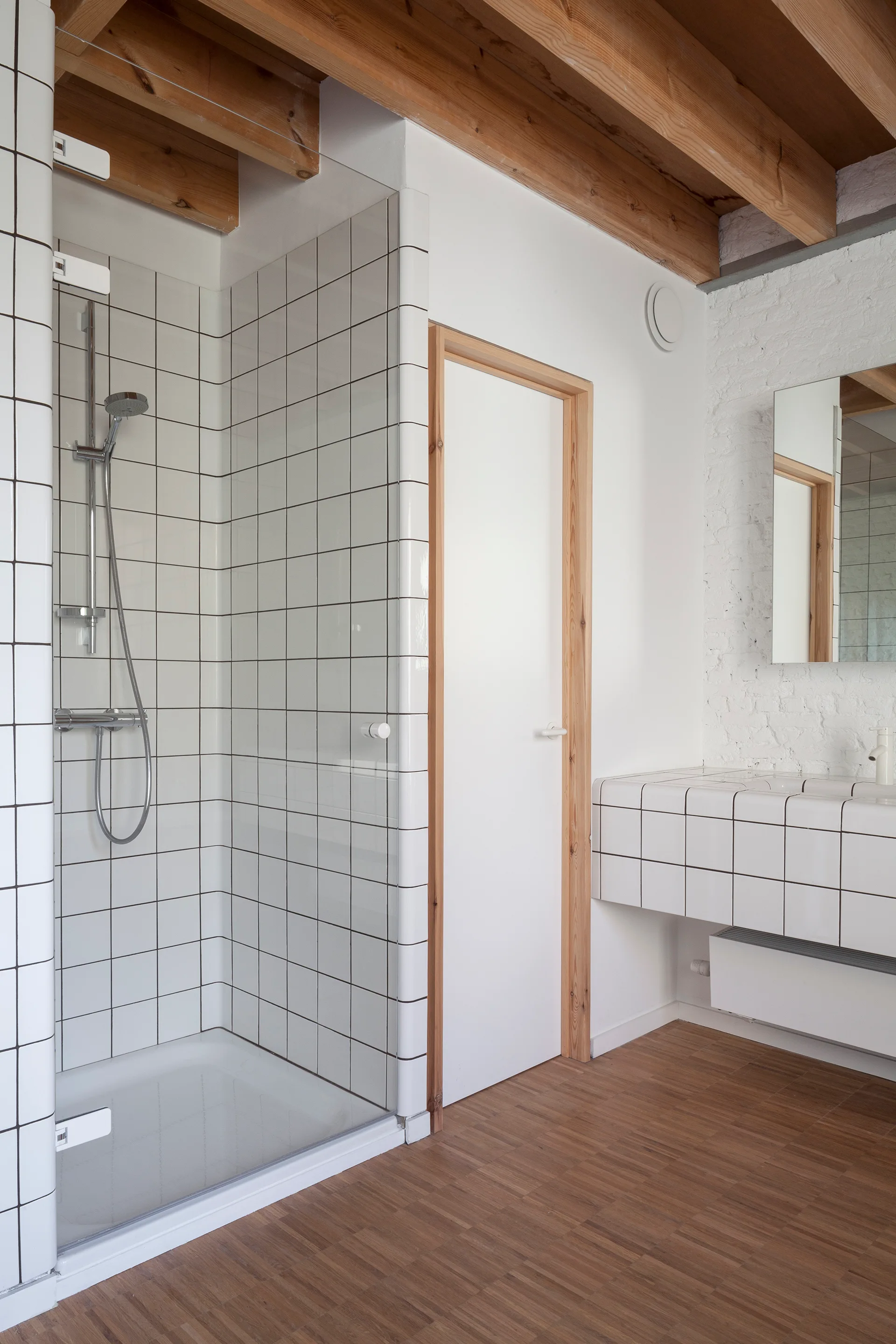 A minimalist bathroom featuring a glass shower stall, a white grid tiled vanity, and exposed wooden ceiling beams.