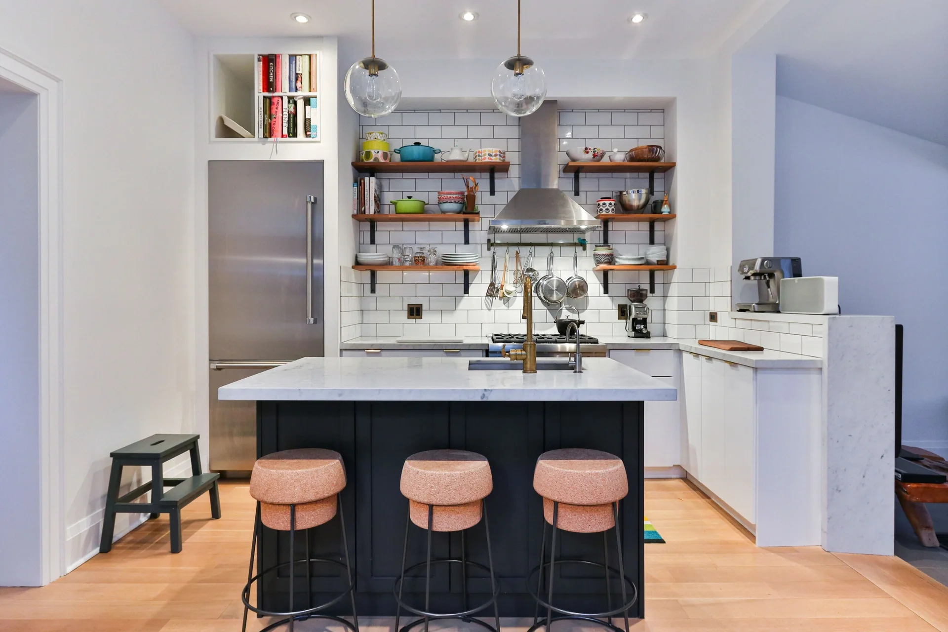 A modern kitchen features a dark blue island with three cork-topped stools, a white subway tile backsplash, and open wood shelves.