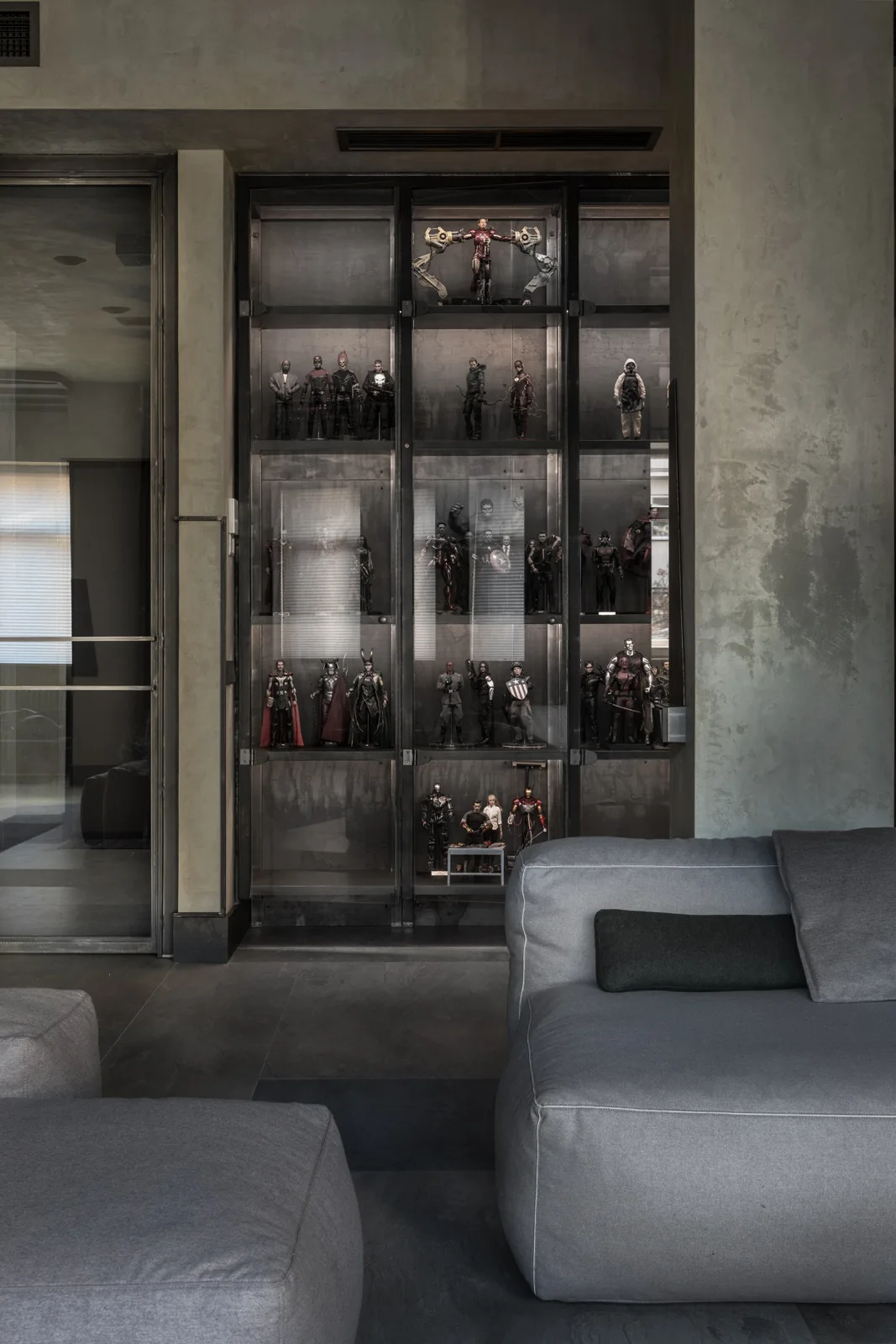 A grey minimalist living room features a built-in illuminated metal display cabinet filled with figurines behind a grey upholstered sofa.