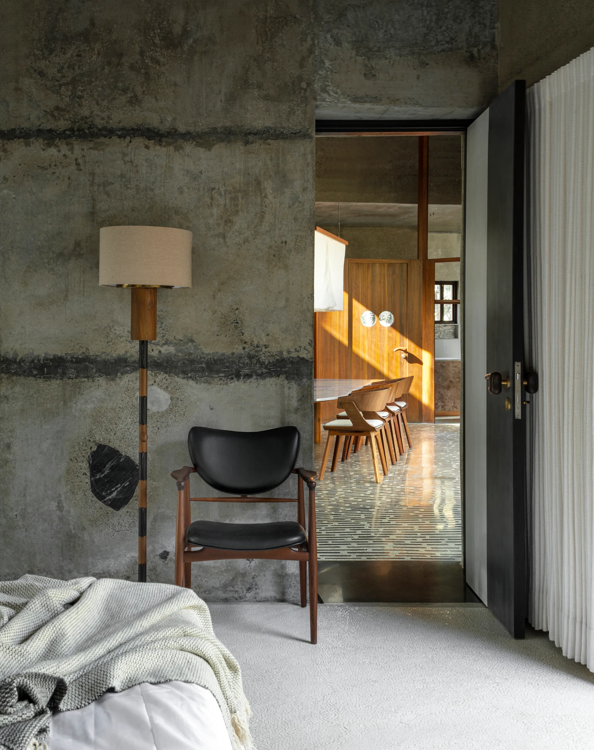 A bedroom with an exposed raw concrete wall features a black leather armchair and a wooden floor lamp next to an open door leading to a dining area.