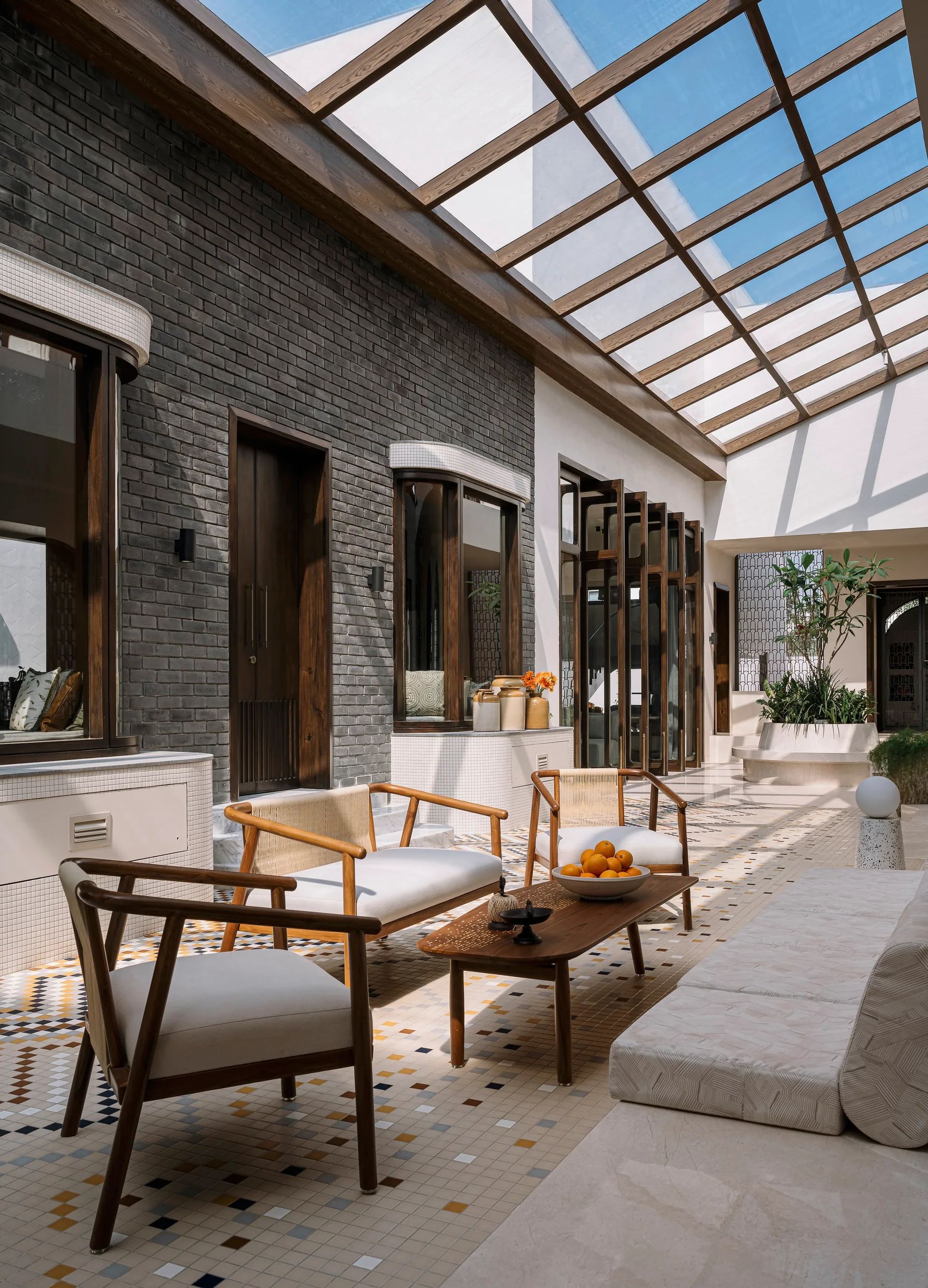 A sunlit courtyard lounge features a glass skylight ceiling, grey brick walls, wood-framed armchairs, and a multi-colored mosaic tile floor.