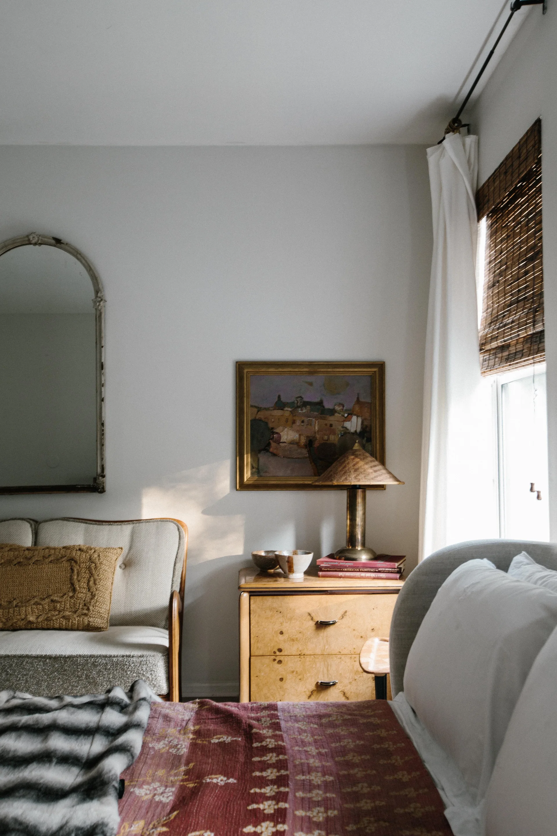 A bedroom corner features a burl wood nightstand, a framed painting, an arched wall mirror, and a small sofa with a mustard yellow throw pillow.