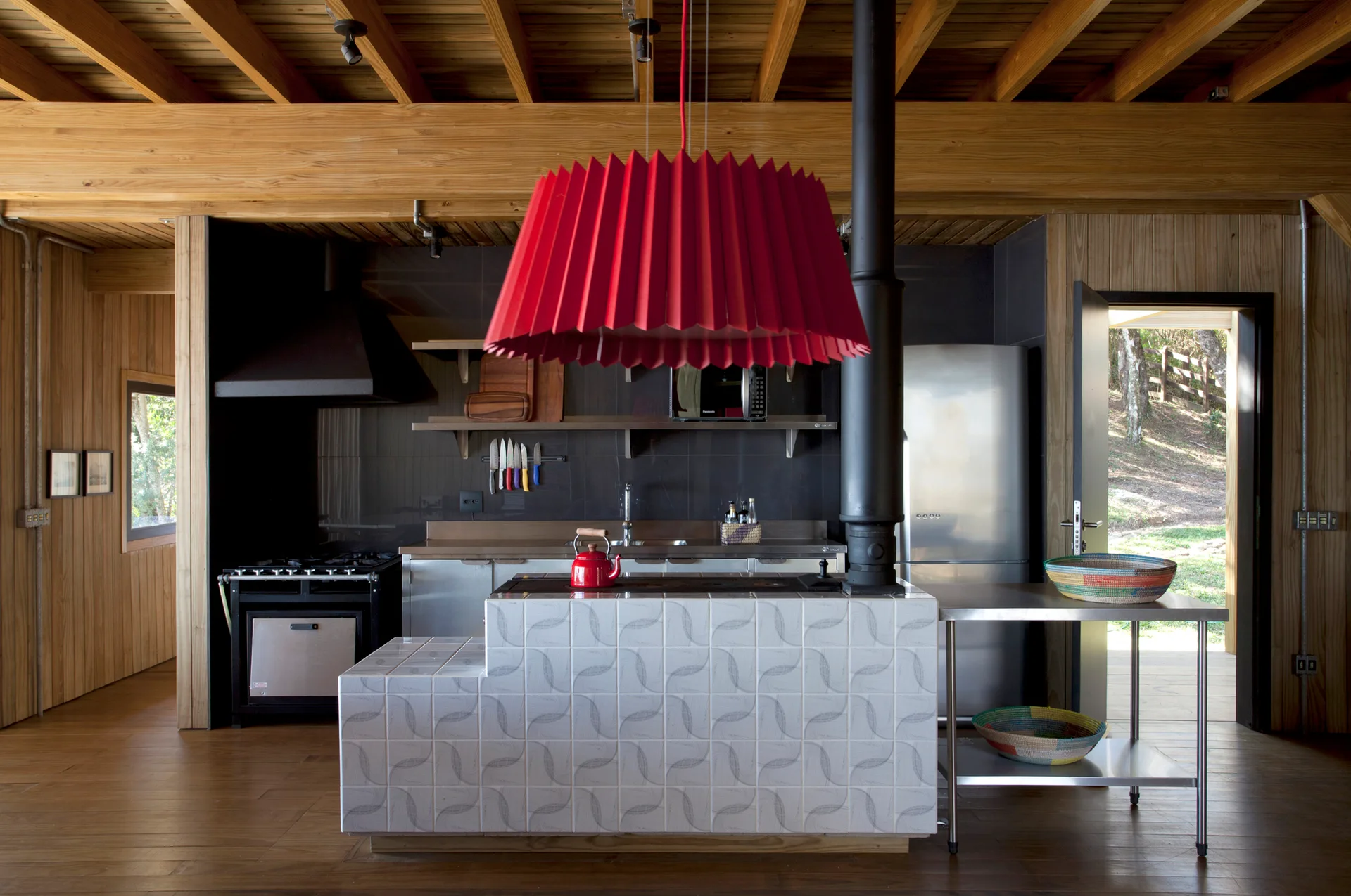 A rustic kitchen features a patterned tile island beneath a large red pleated pendant light, surrounded by wood paneled walls and ceilings.