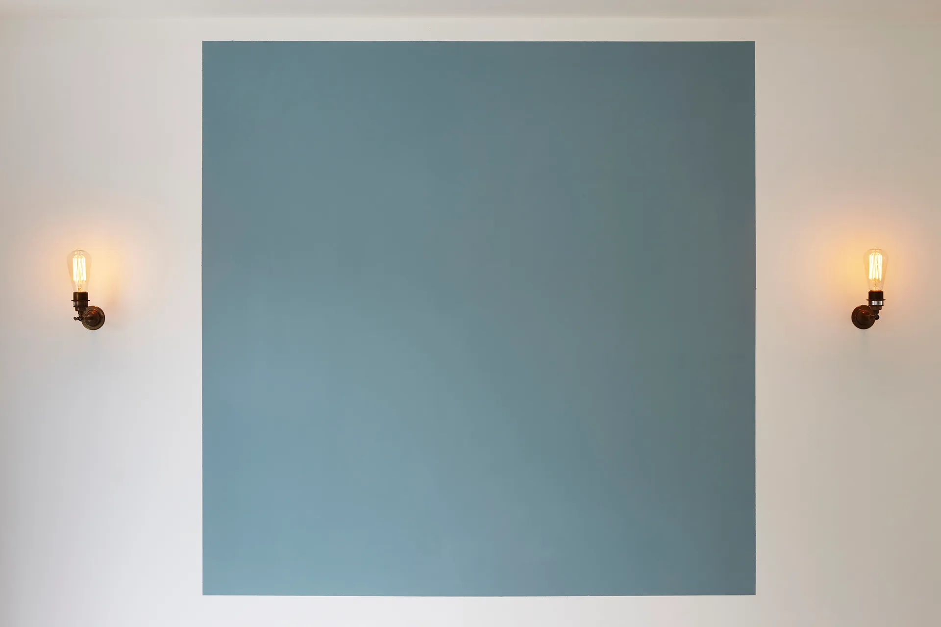 A large blue square painted on an off-white wall is centered between two glowing metal sconces with vintage Edison bulbs.