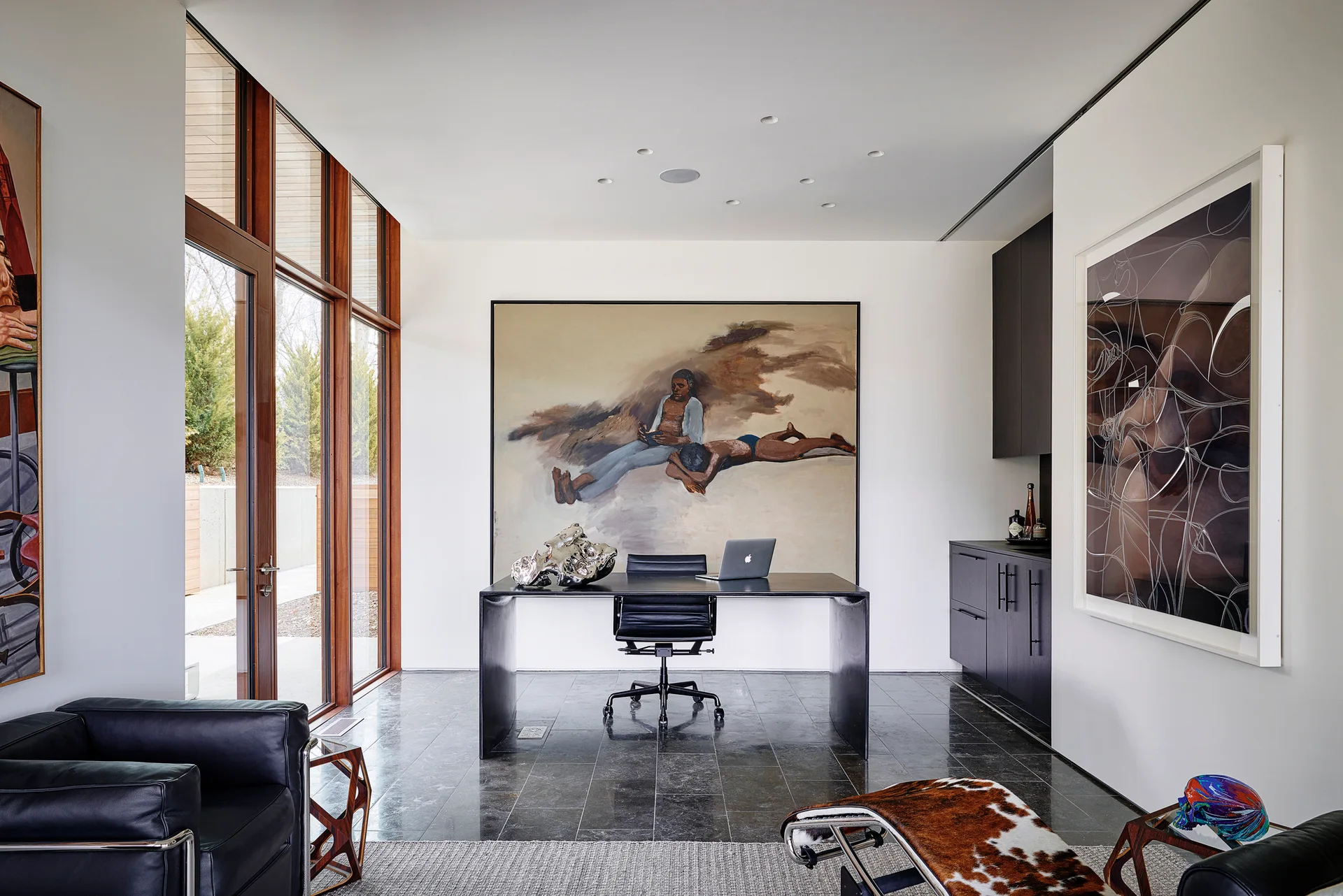 A modern home office features a black desk and chair, a large painting on the wall, dark stone tile floors, and floor-to-ceiling windows.