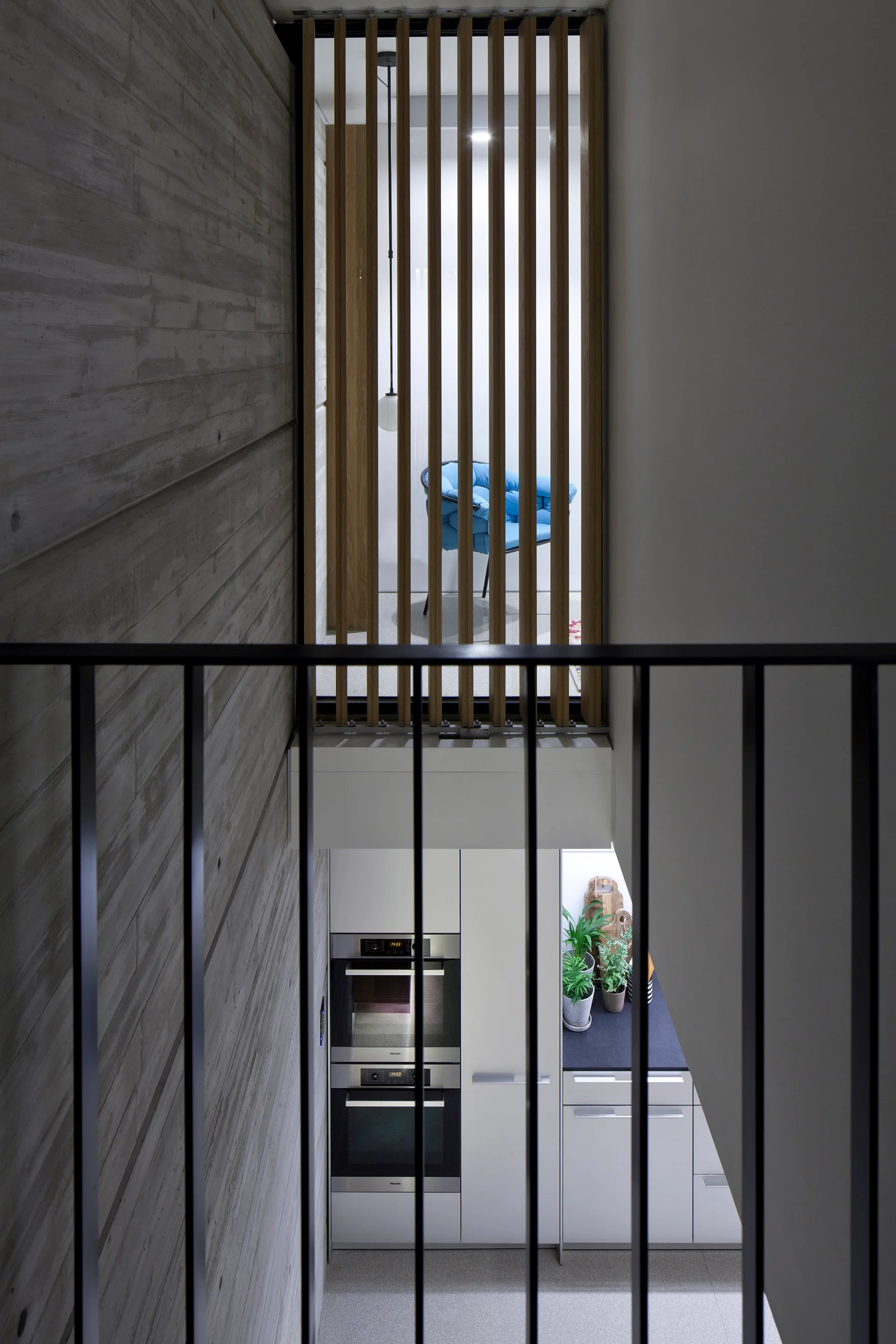 A view down a modern staircase shows a board-formed concrete wall, a black metal railing, a vertical wood slatted screen, and a kitchen below.