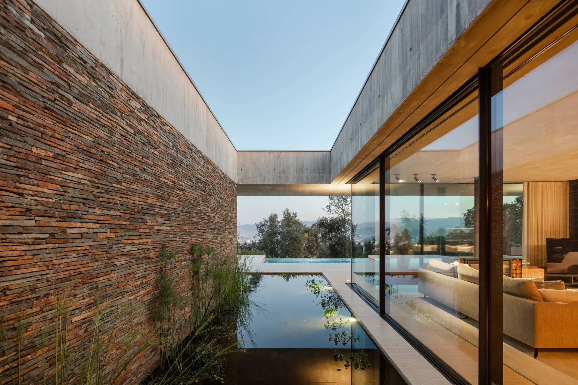 A modern outdoor courtyard features a reflection pool, a textured stacked stone wall, and a large glass window looking into an indoor living room.
