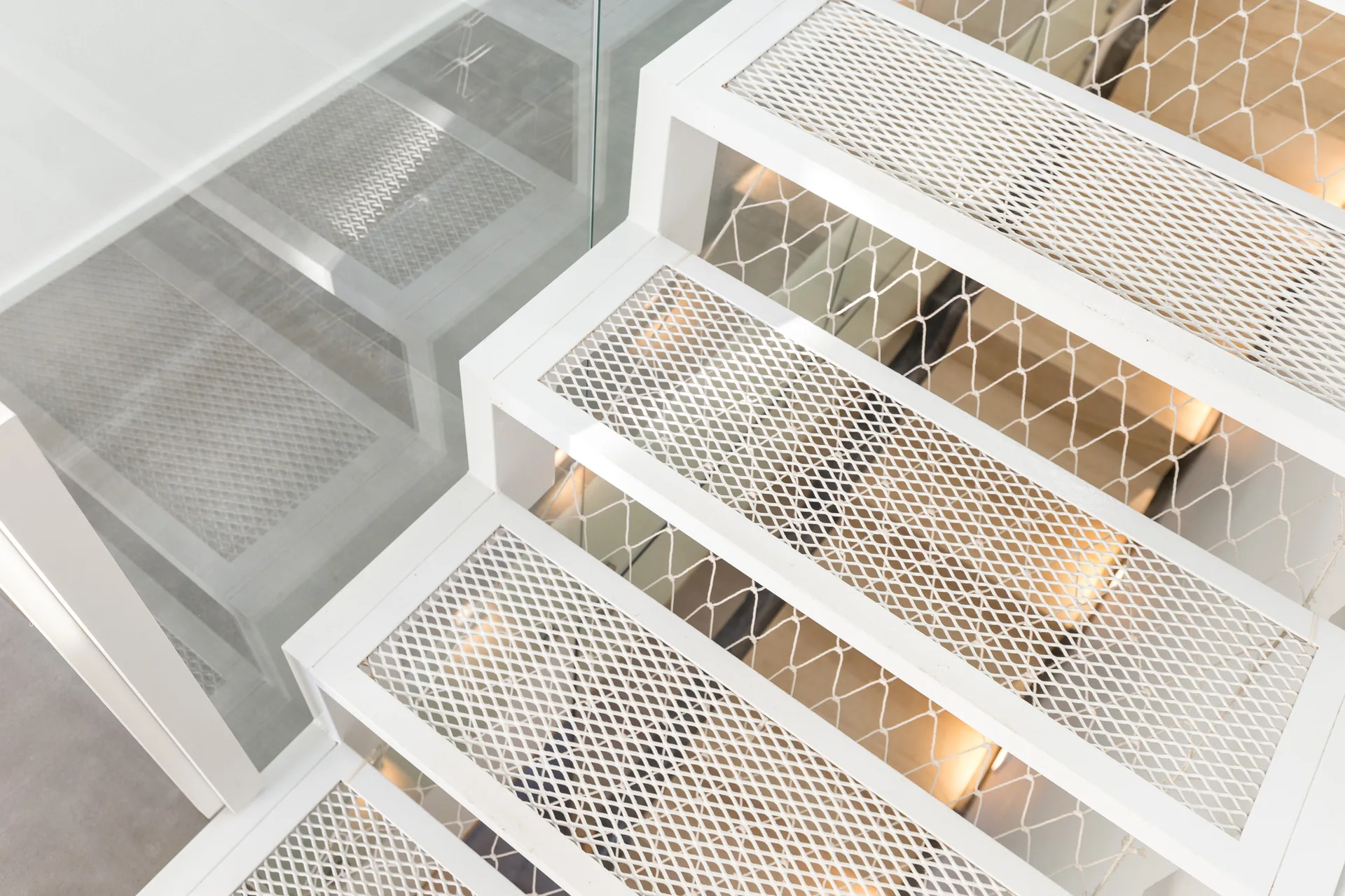 A close-up, high-angle view of a modern staircase with white metal mesh treads, a clear glass railing, and decorative safety netting.