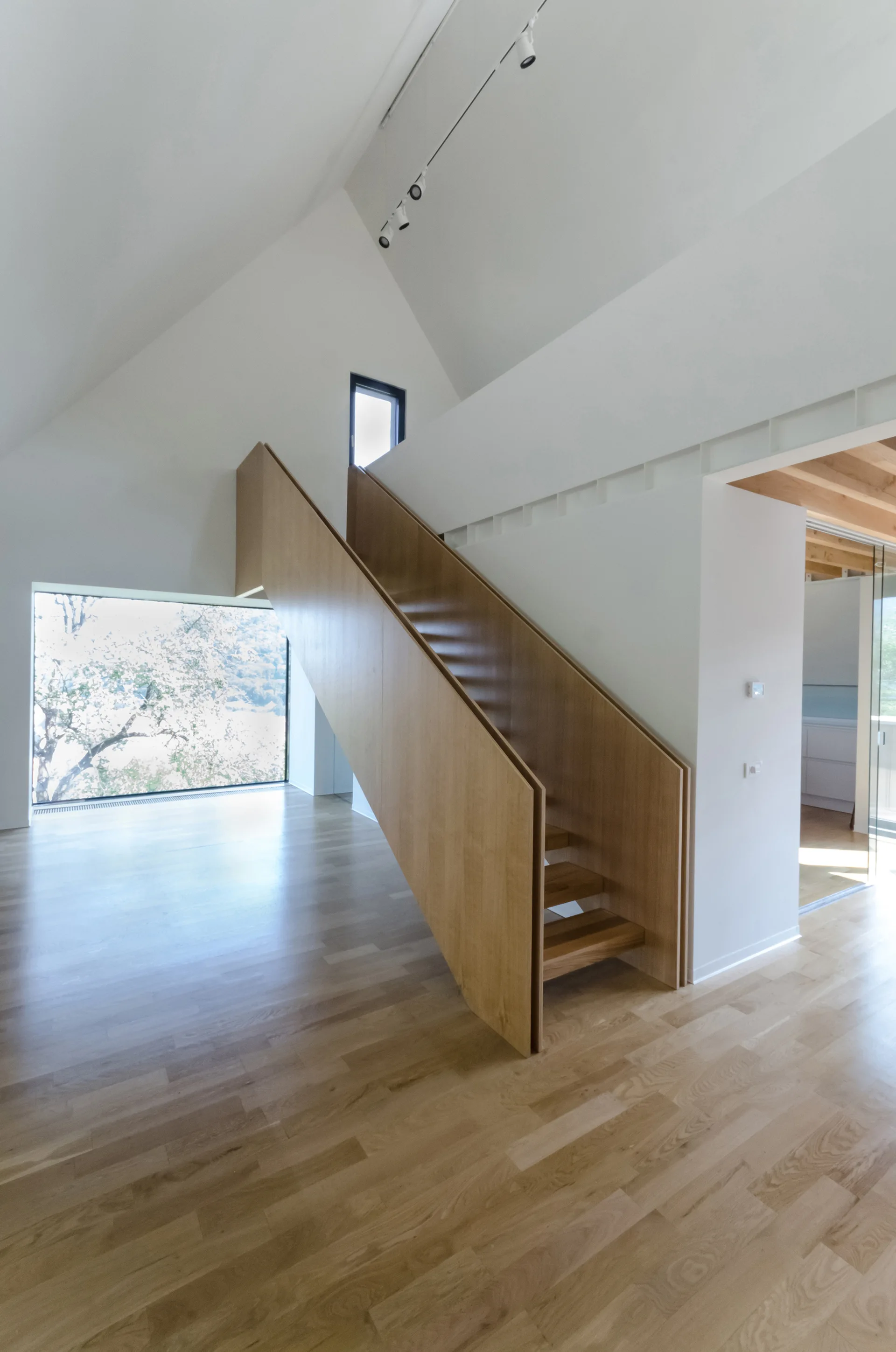 A modern minimalist staircase with solid light wood side panels sits in a high-ceilinged white room with matching wood floors and a large window.