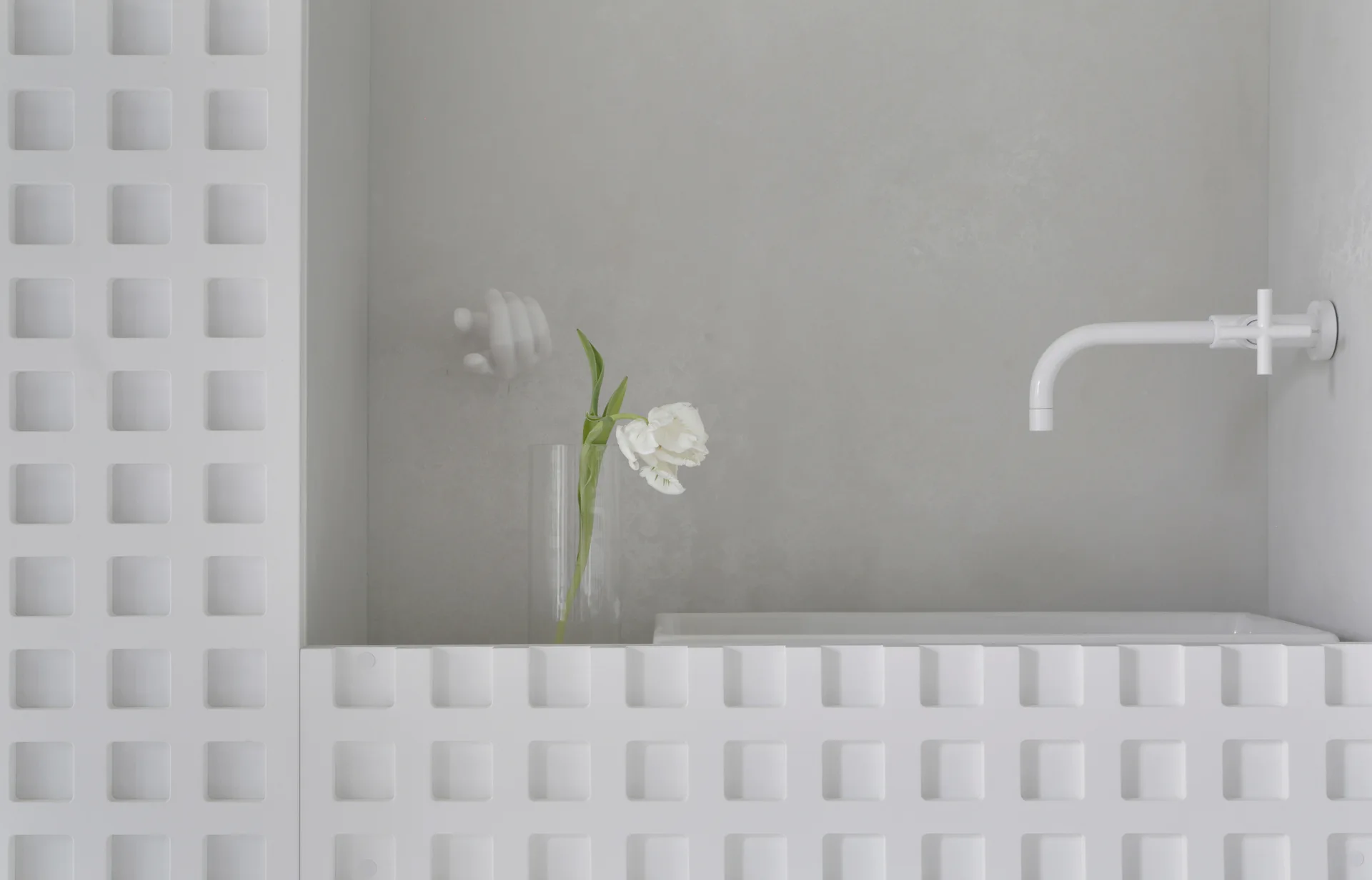 A minimalist bathroom sink features a white wall-mounted faucet, a grid-textured wall panel, and a single white tulip in a clear glass vase.