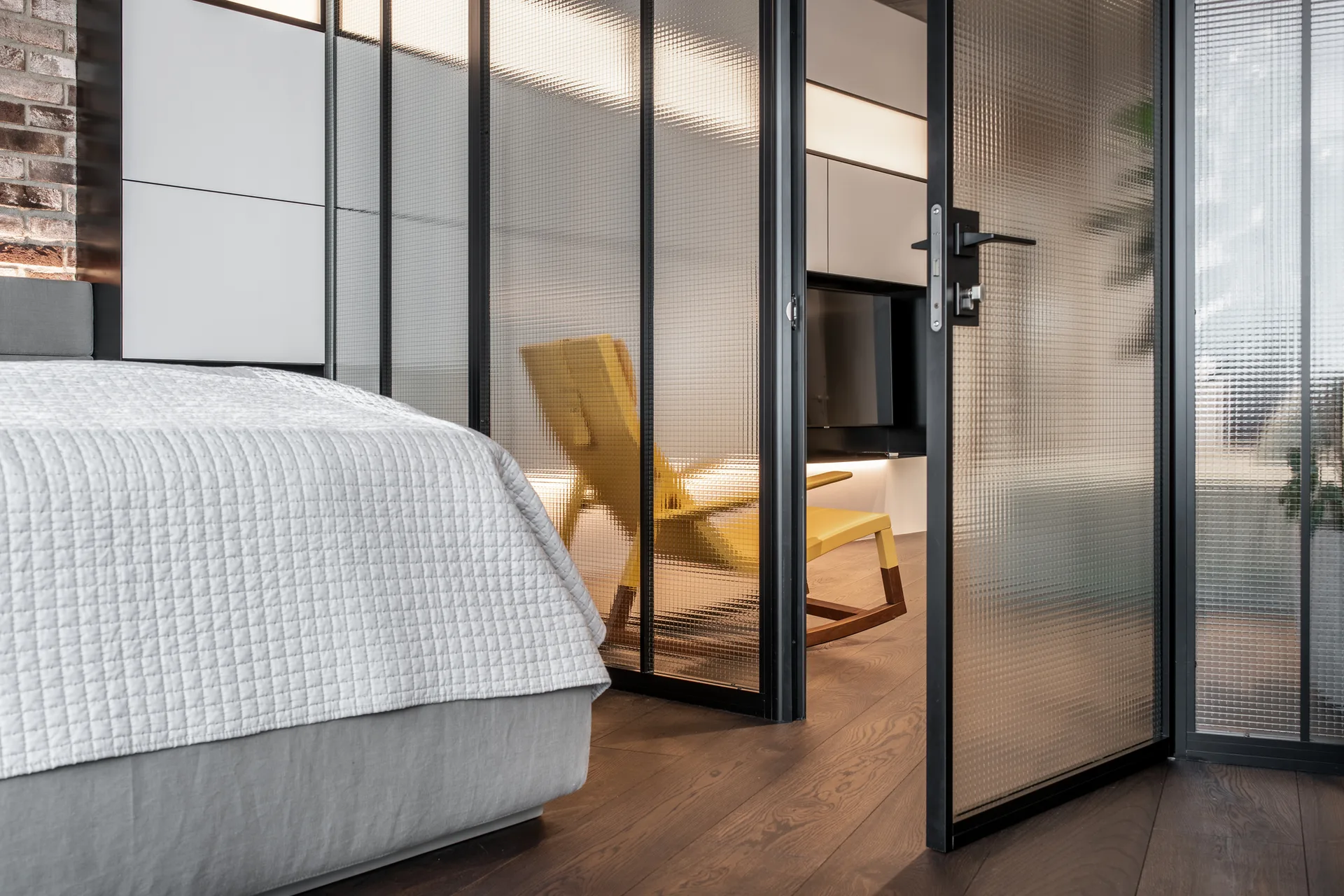A contemporary bedroom with a grey bed is separated from an adjacent room with a yellow chair by black-framed textured glass panels.