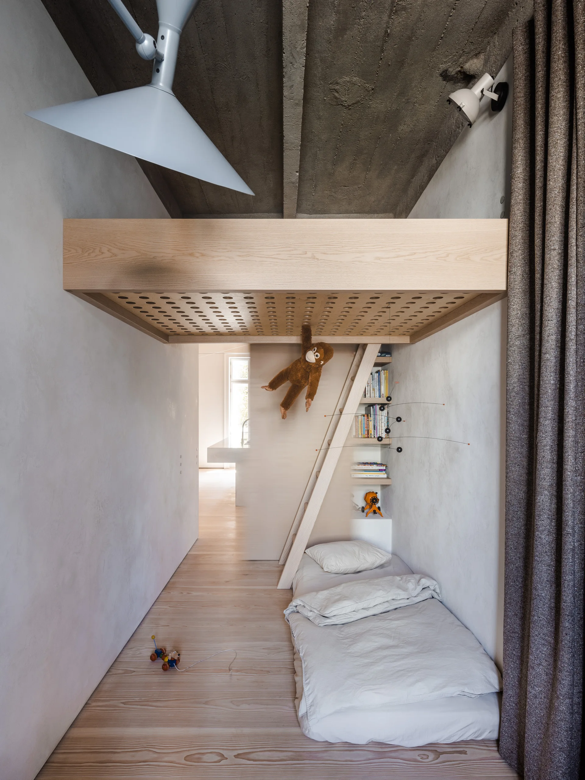 A minimalist kids' bedroom features a light wood loft bed with a ladder, a floor mattress, plaster walls, and an exposed raw concrete ceiling.