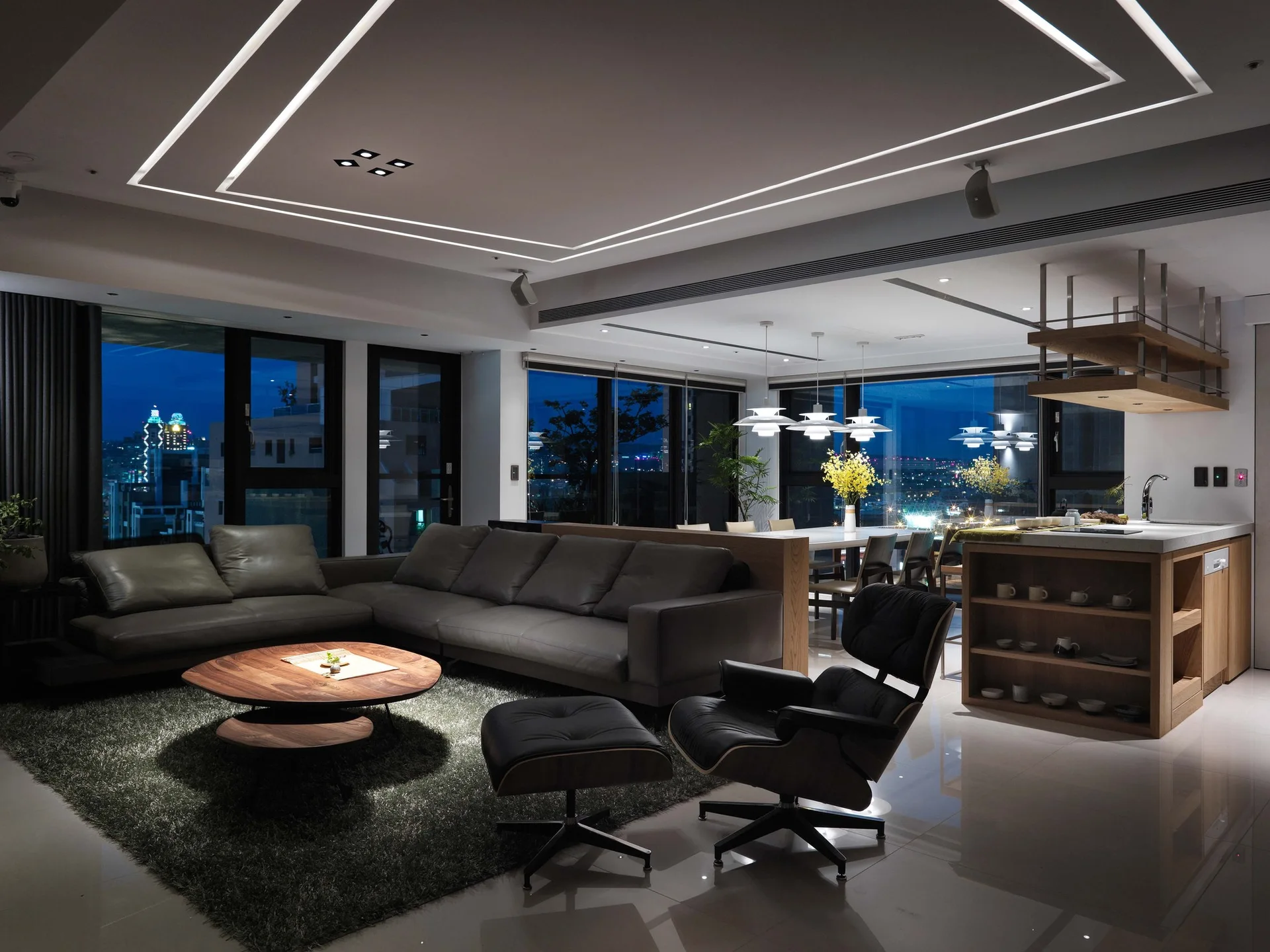 A modern open-plan living room features a grey leather sectional, a wooden coffee table, and a kitchen island against windows with a night city view.
