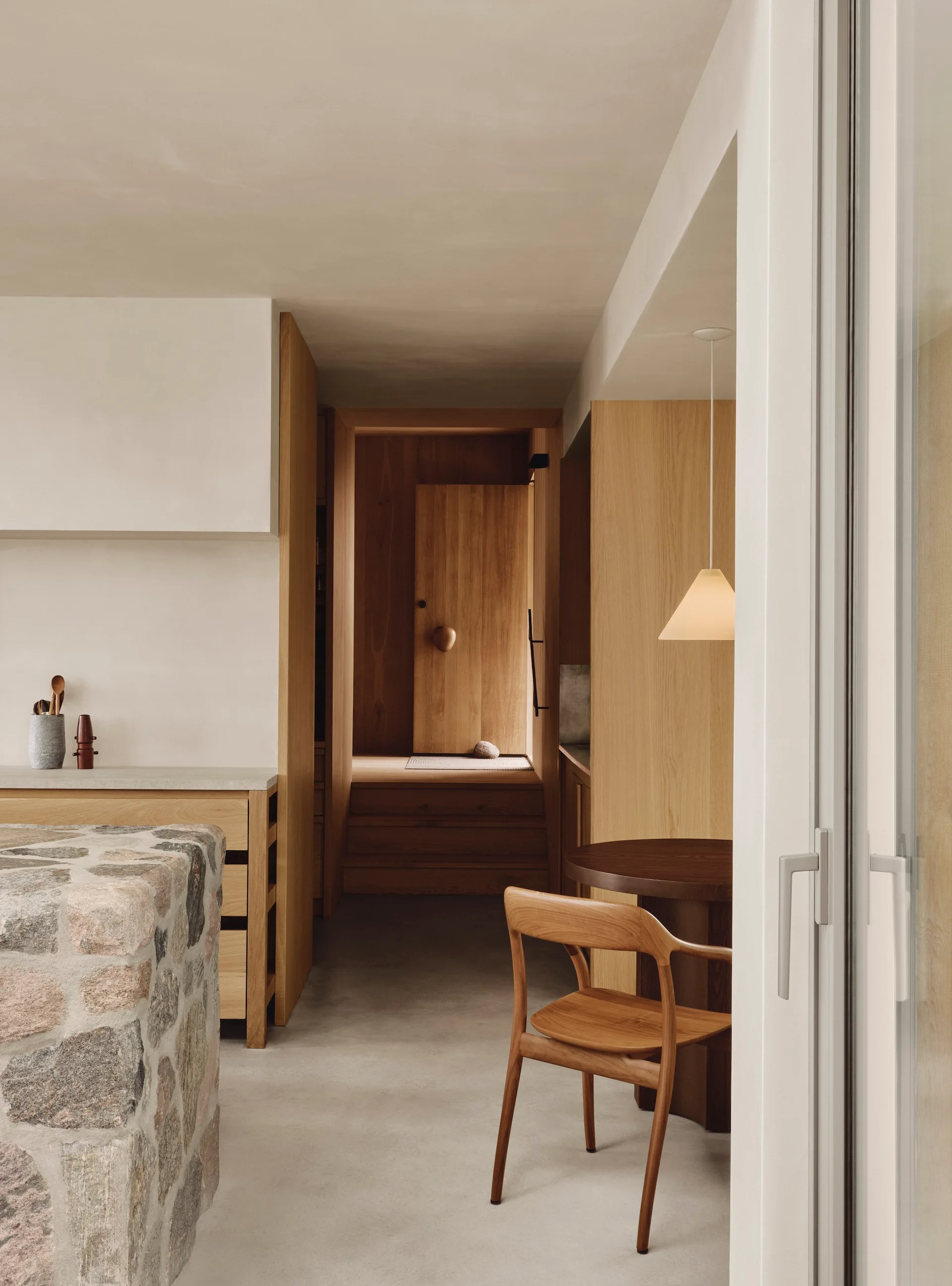 A minimalist kitchen view showing a textured stone island, warm wood paneling, a dining table with a wooden chair, and a glowing pendant light.