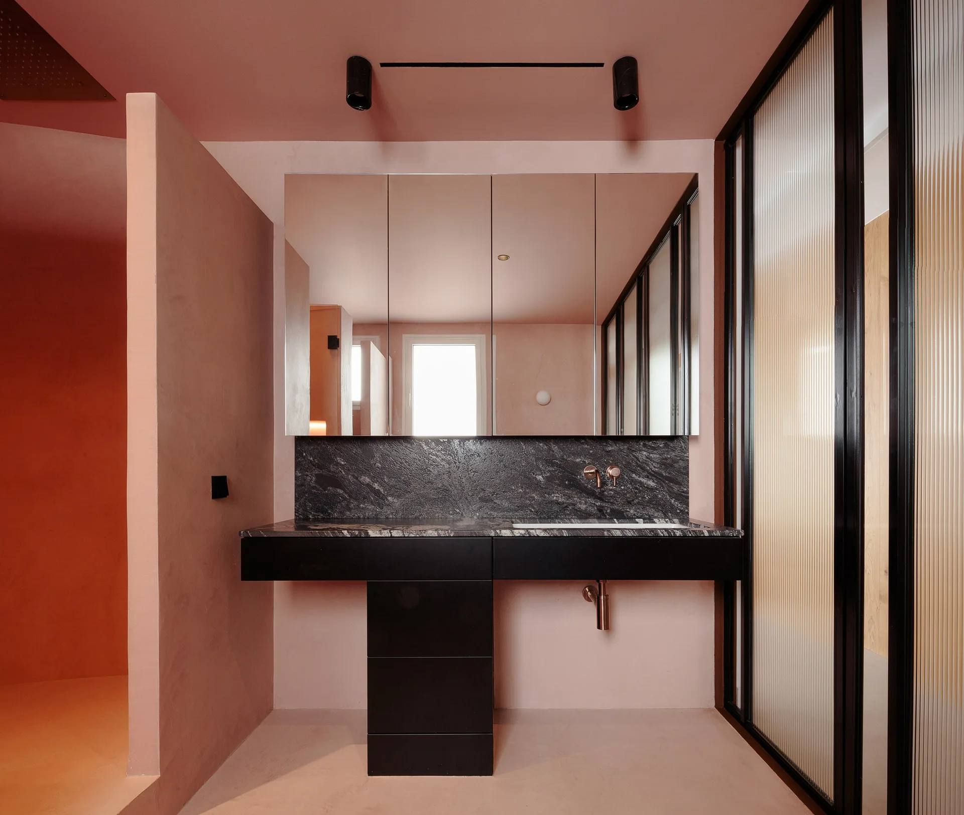 A minimalist bathroom featuring a black stone vanity against pink walls and ceiling, flanked by a fluted glass partition with a black metal frame.
