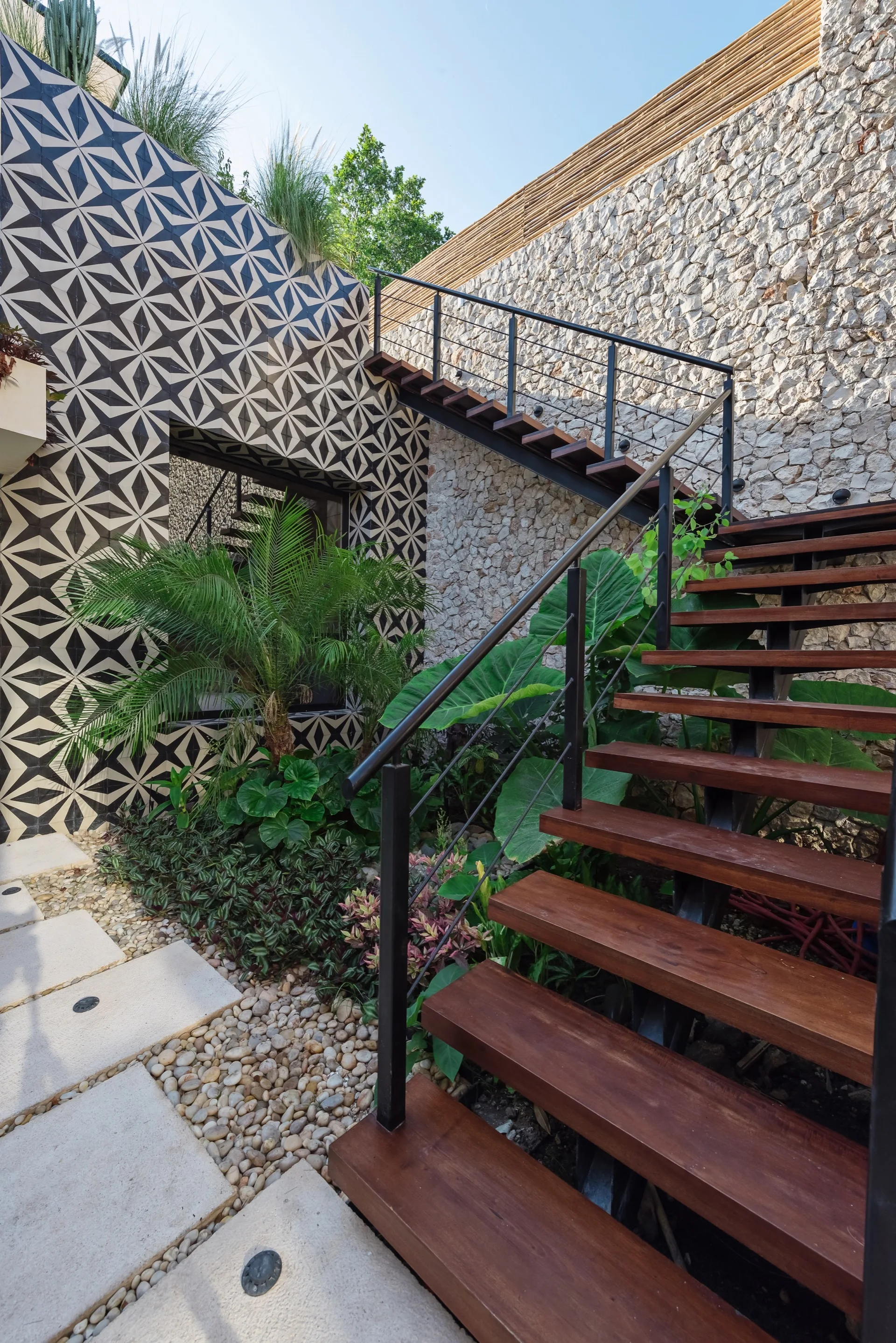 Floating wooden stairs rise past a rough stone wall and a bold geometric tiled wall in a landscaped outdoor patio with tropical plants.