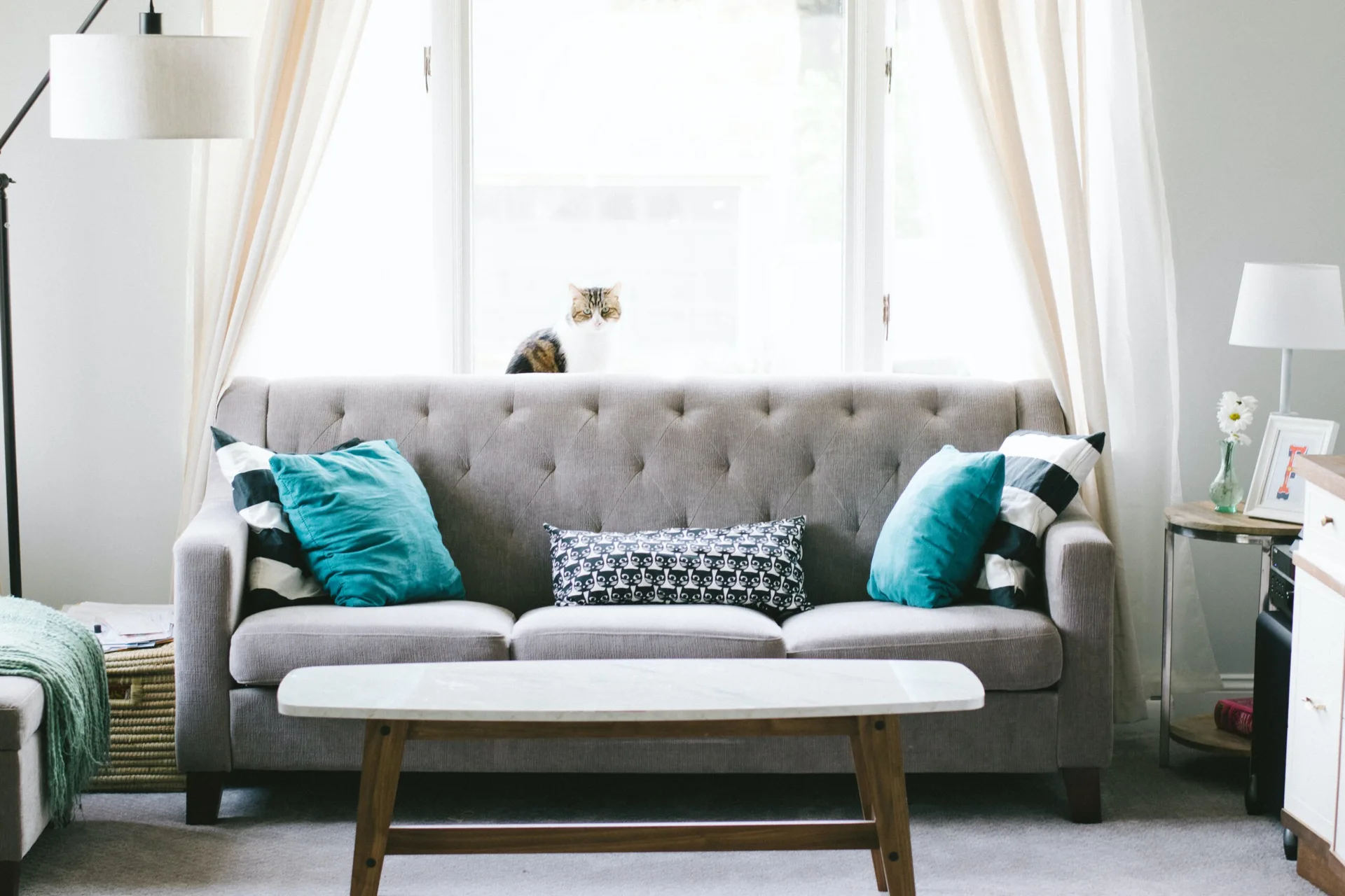 A light grey tufted sofa with teal and patterned pillows sits in front of a window with white curtains, accompanied by a marble-top coffee table.