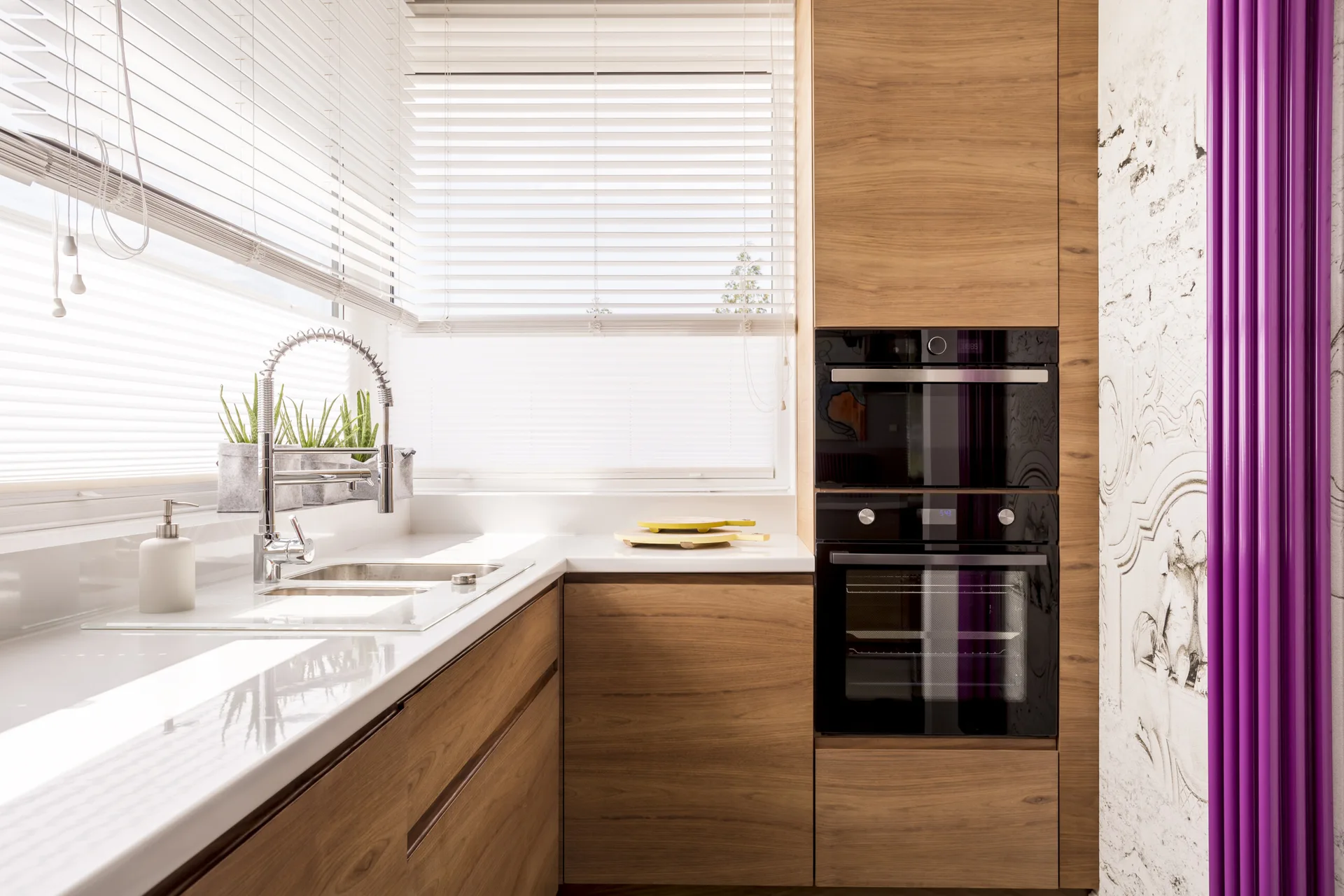 A modern kitchen corner with warm wood-toned cabinets, a stainless steel sink under white window blinds, and a vibrant purple vertical radiator.