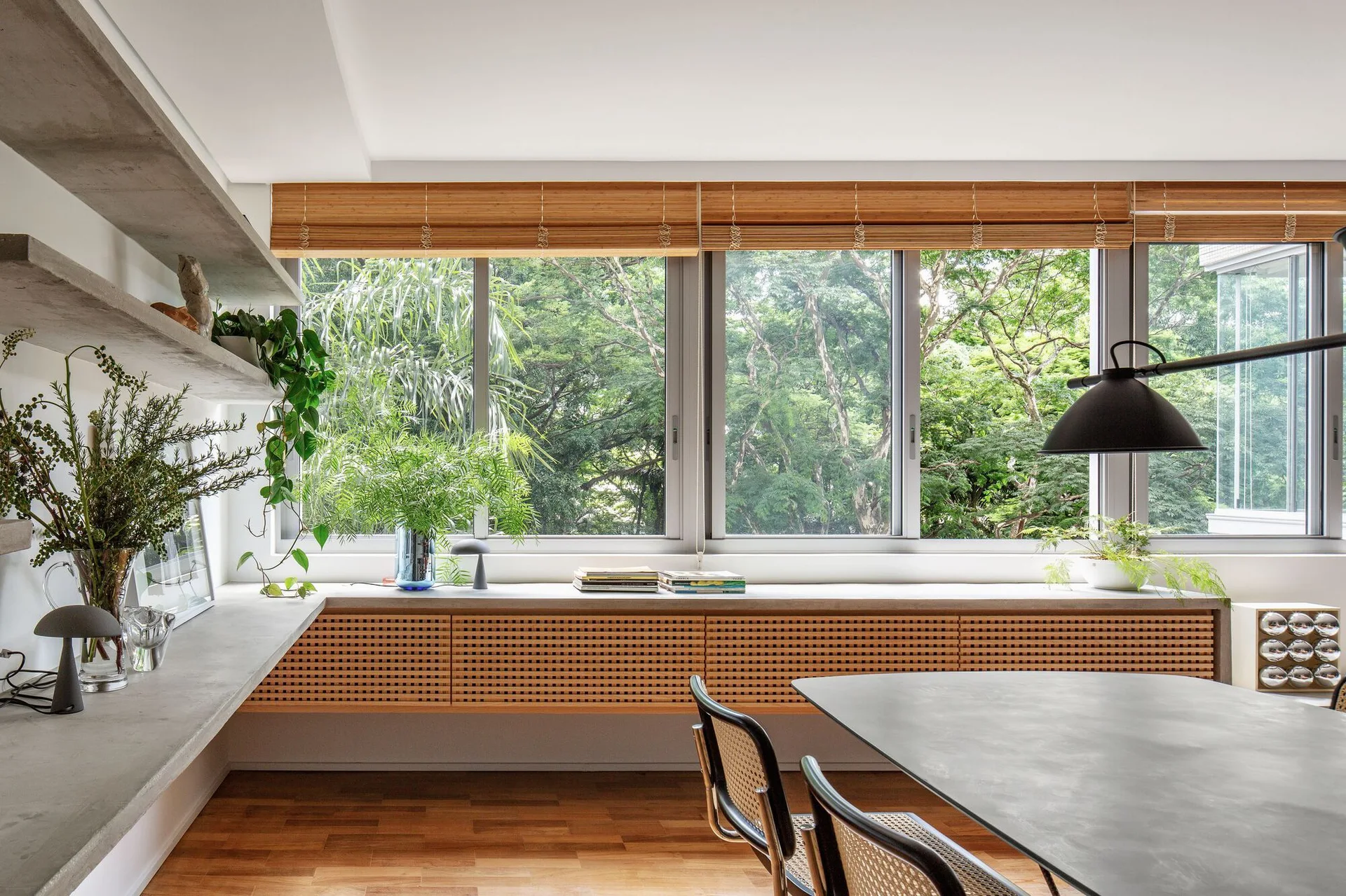 A modern dining room features a dark table, cane-backed chairs, a concrete floating shelf, and a low console beneath wide windows with bamboo blinds.