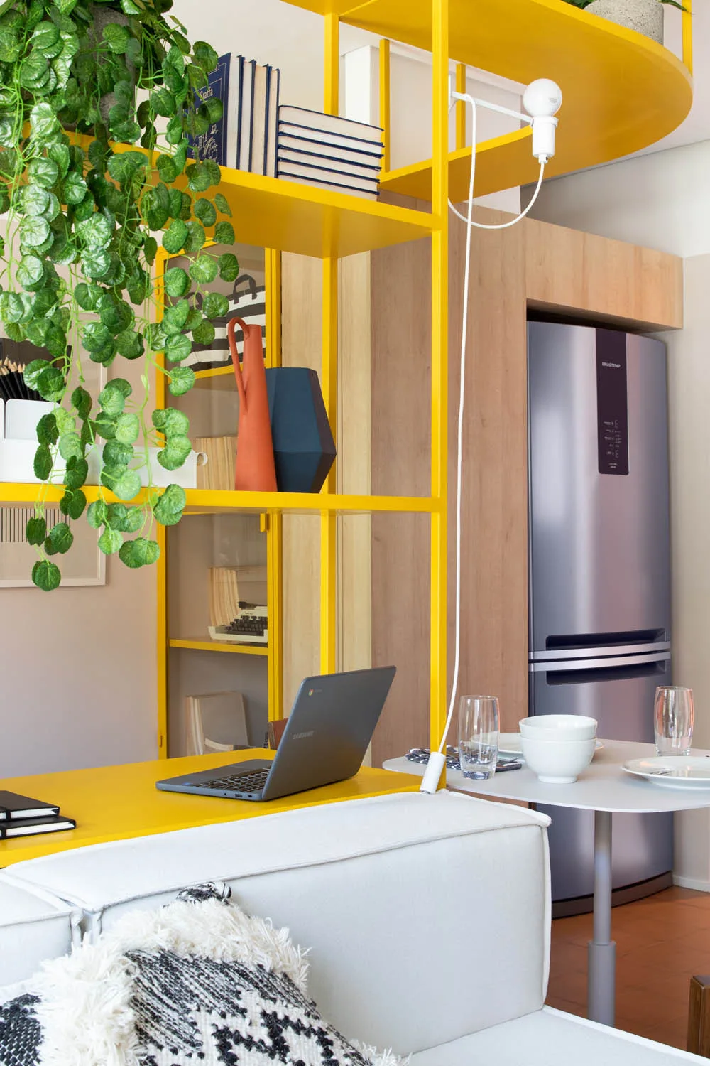 A bright yellow open shelving unit with books and plants separates a white sofa in the foreground from a kitchen area with a refrigerator.