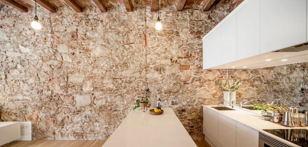 Sleek white kitchen cabinets and a matching island contrast with a massive rustic stone wall and exposed wood ceiling beams.