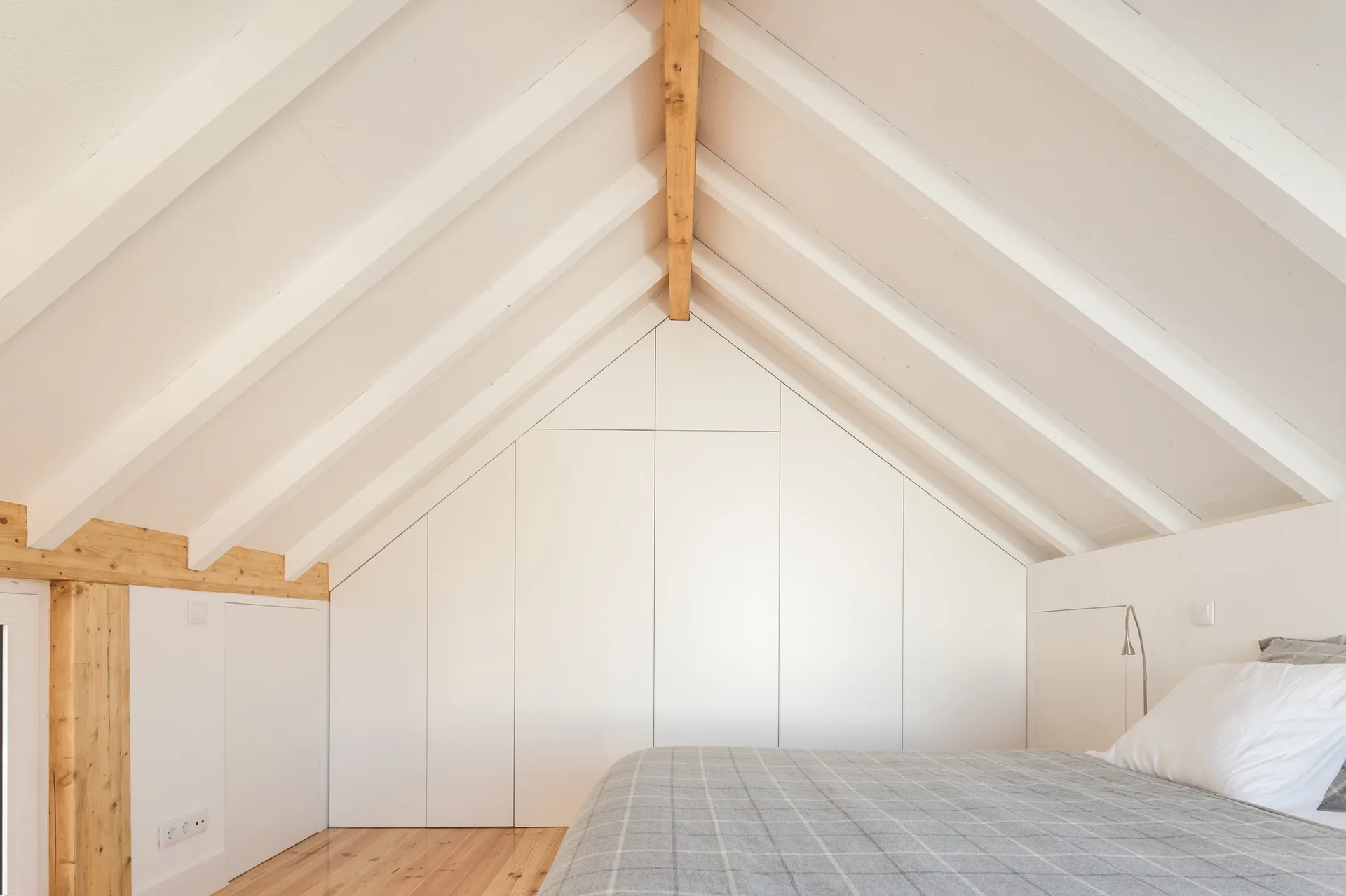 An attic bedroom features white custom wardrobes tailored to the sloped ceiling, exposed wood rafters, and a bed with a grey plaid cover.