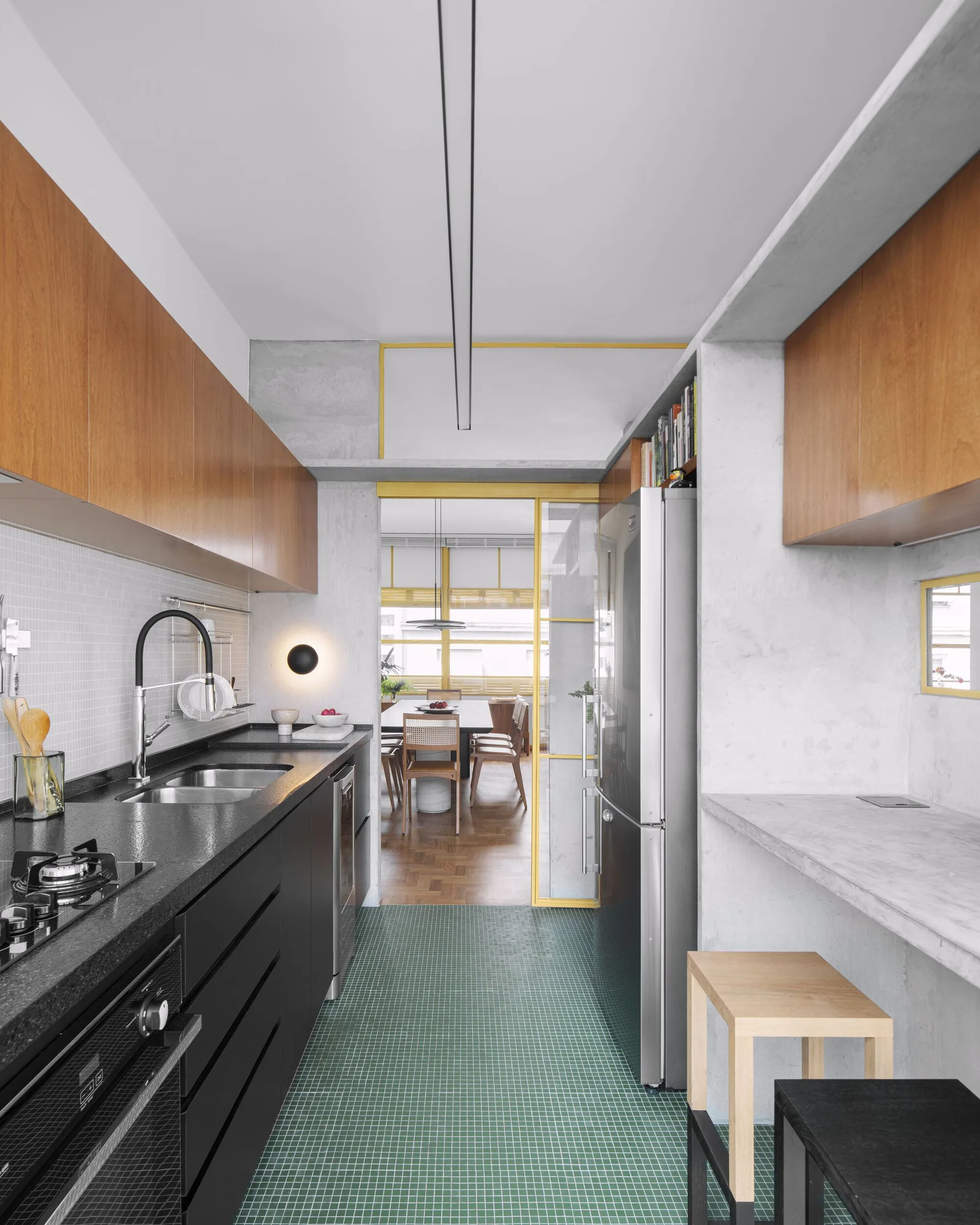 A modern galley kitchen featuring green tiled floors, black lower cabinets, raw concrete walls, and a yellow-framed sliding glass door.