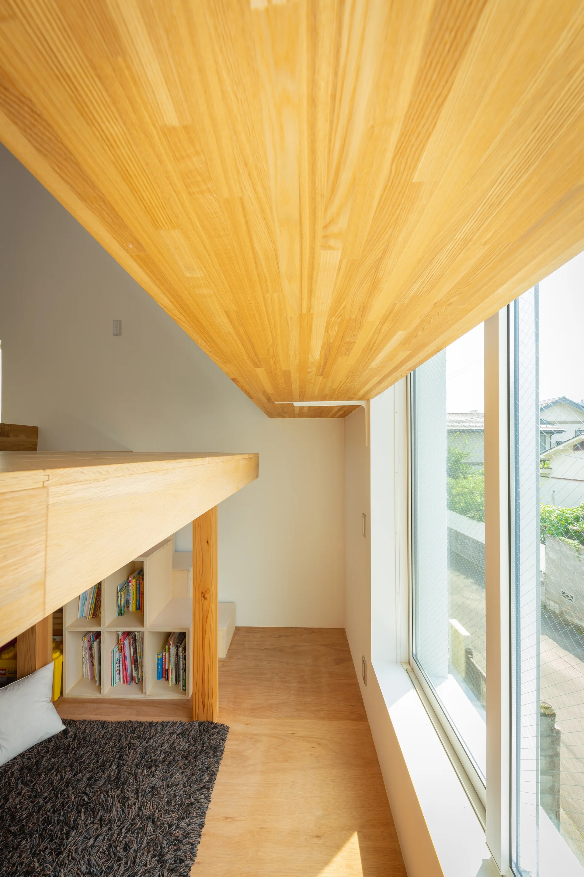 A minimalist wooden loft platform features built-in book cubbies underneath and a large window looking out onto a residential area.