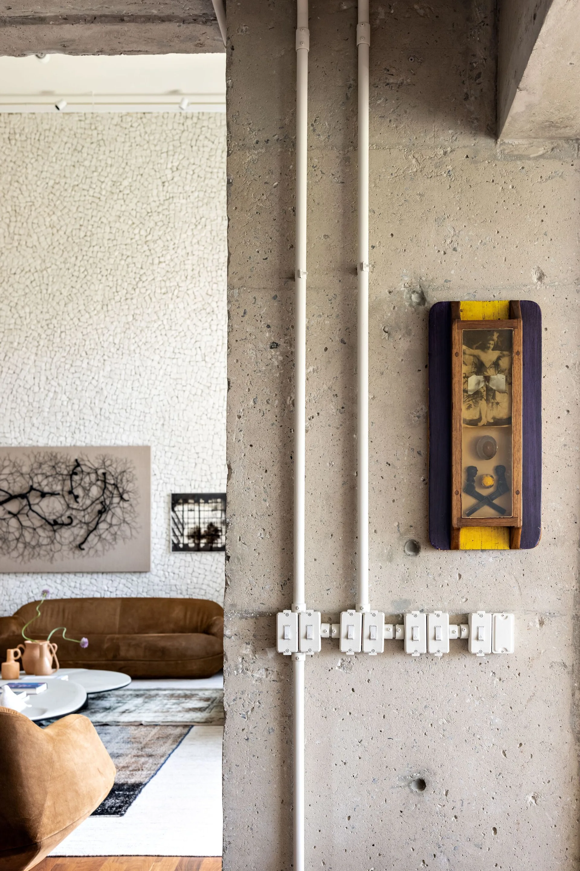 A raw concrete column with white conduits and light switches frames a view into a living room with a brown sofa, round coffee table, and textured wall.