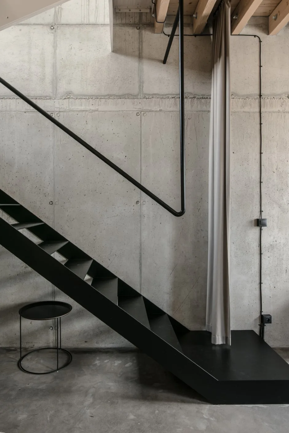 A black metal staircase rises against a gray concrete wall next to a hanging beige curtain and a round metal side table.
