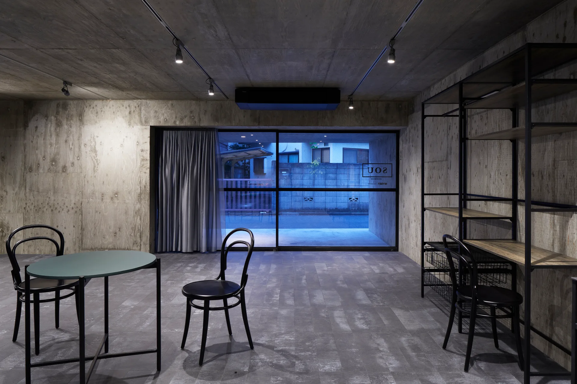 An industrial room with raw concrete walls features black bentwood chairs, a green-topped round table, and a tall metal shelving unit.