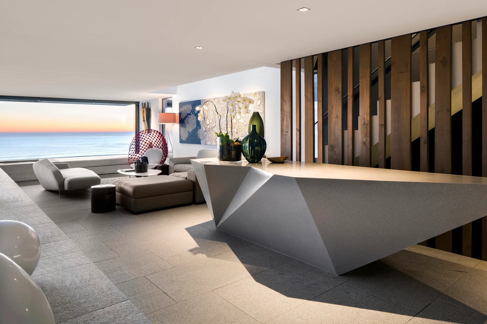 A modern coastal living room with a large faceted gray console table, vertical wood slat screen partition, and an ocean view at sunset.