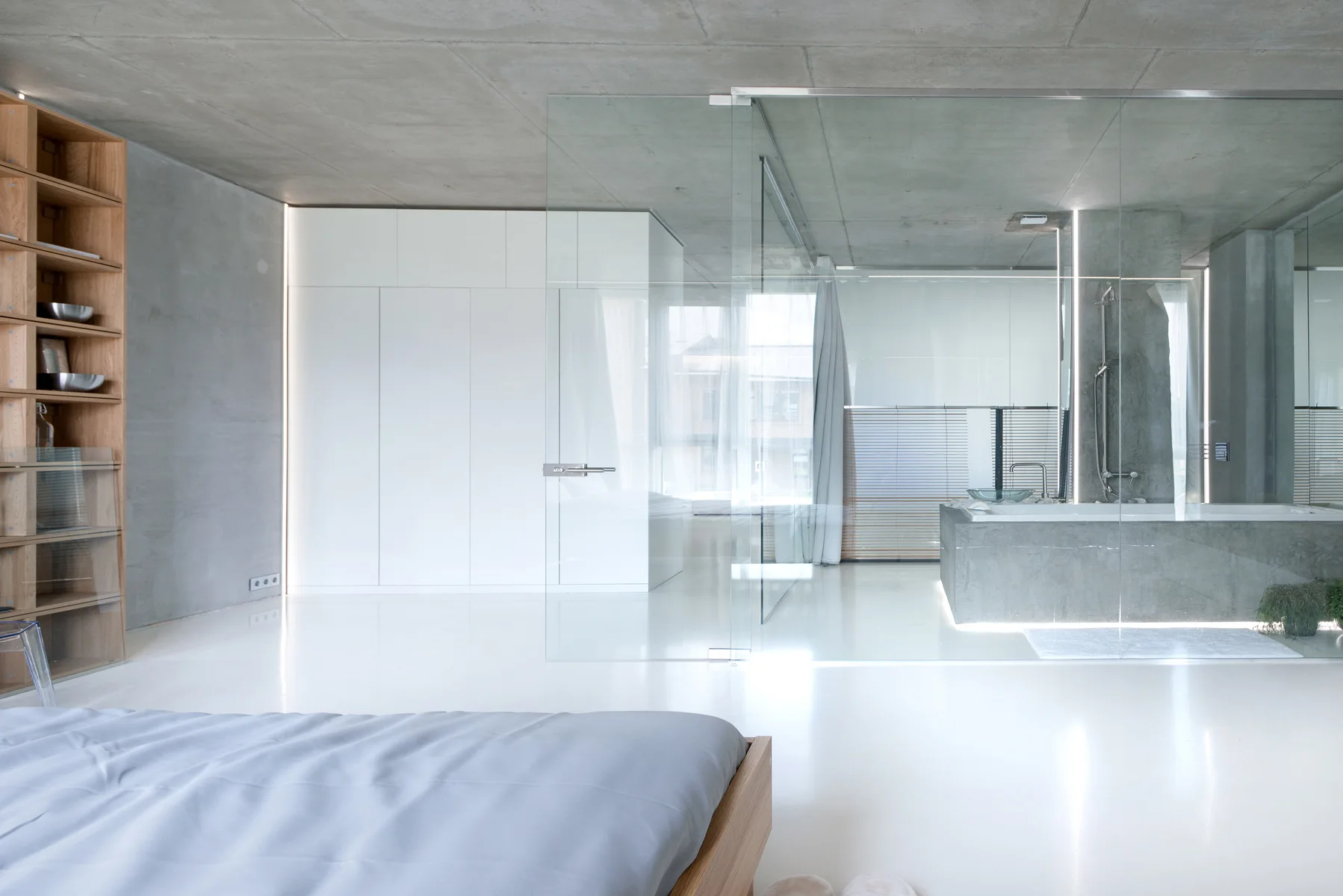 A minimalist open-plan bedroom featuring a glossy white floor, exposed concrete ceiling, and a glass-enclosed bathroom area.
