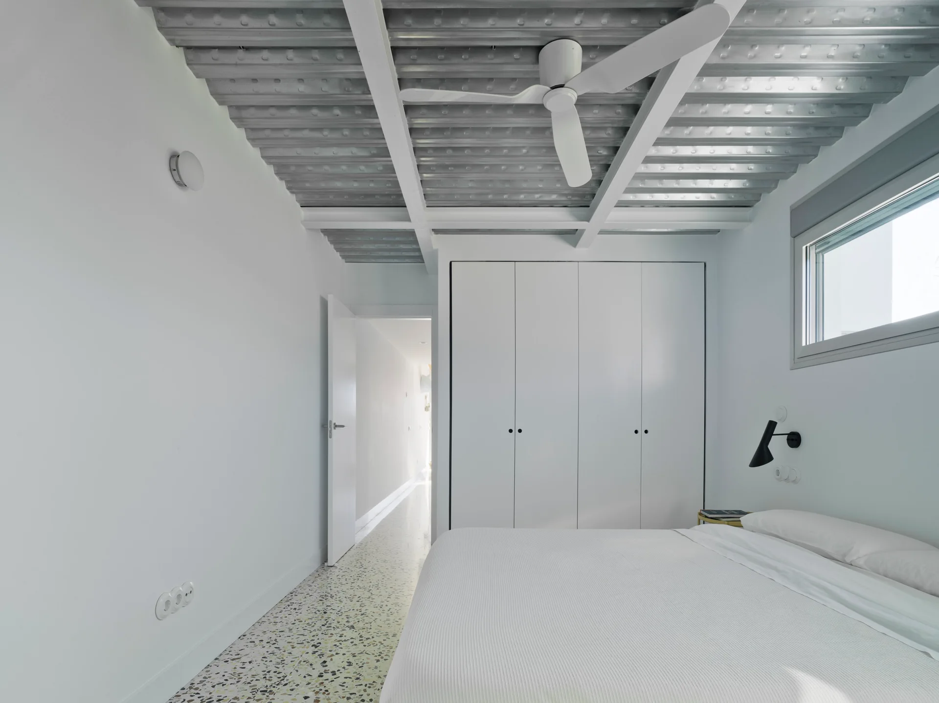 A modern white bedroom featuring an exposed corrugated metal ceiling with white beams, terrazzo flooring, and a built-in wardrobe.