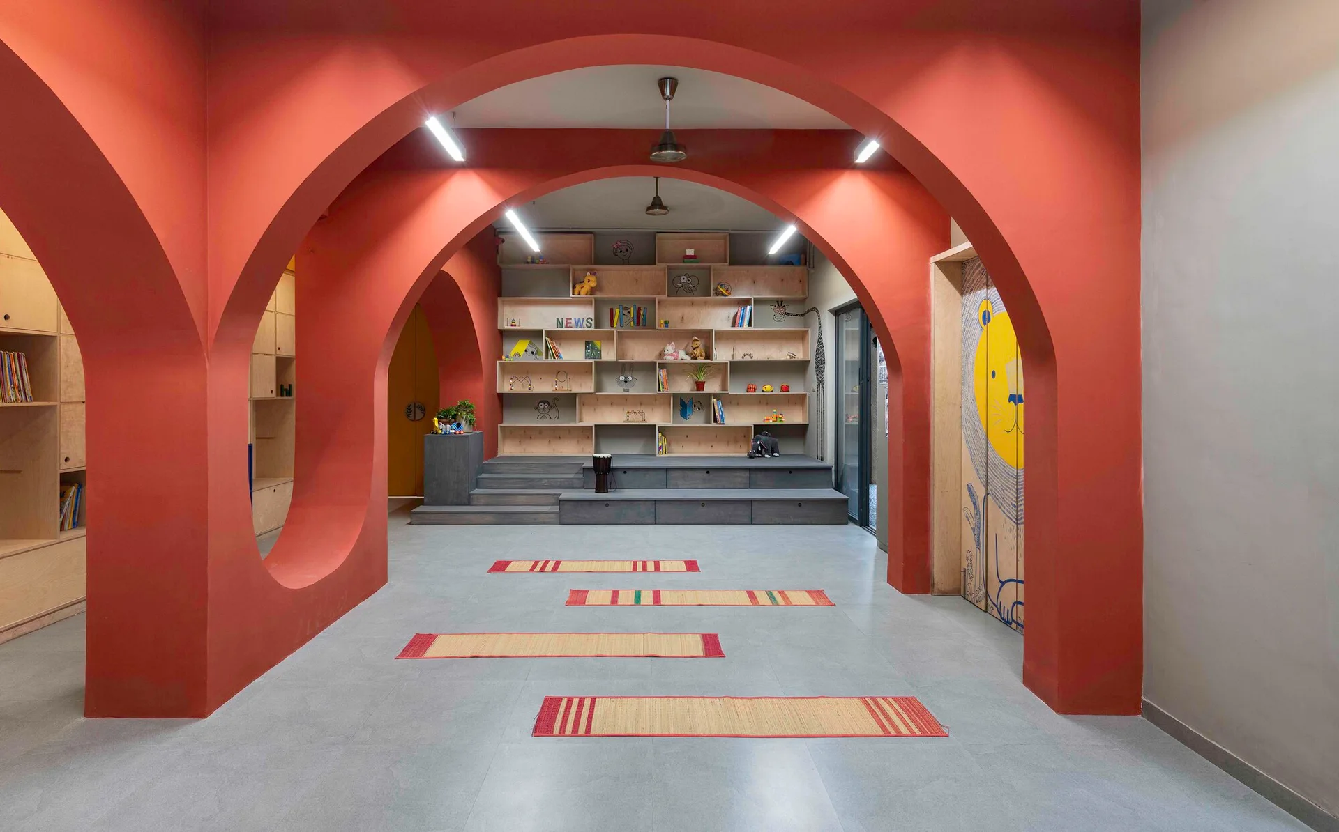 A spacious playroom features bold red arches, a large light-colored plywood shelving unit with a built-in platform stage, and four thin floor mats.