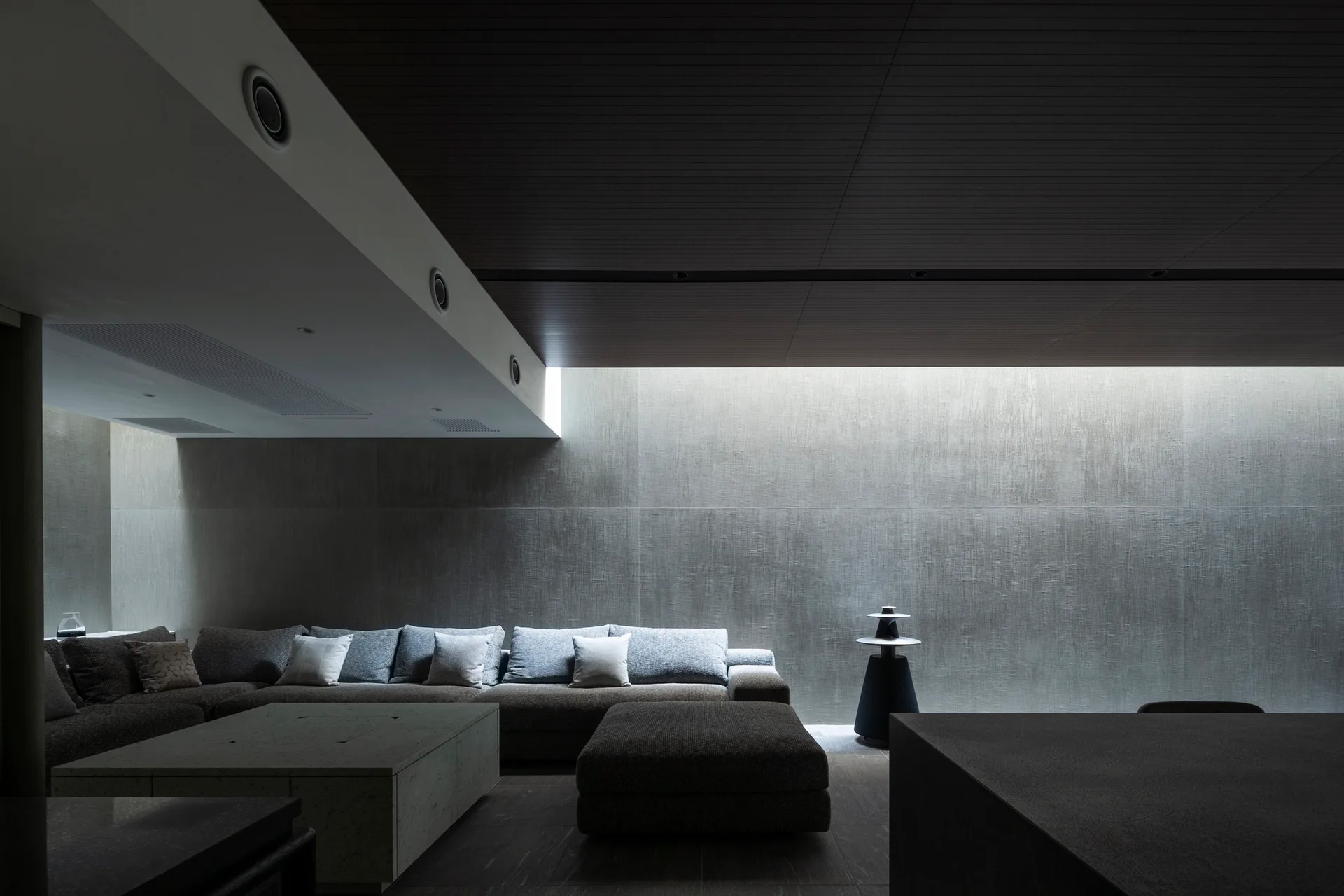 A minimalist living room features a grey sectional sofa, a stone coffee table, and an accent wall washed in soft cove lighting under a dark ceiling.