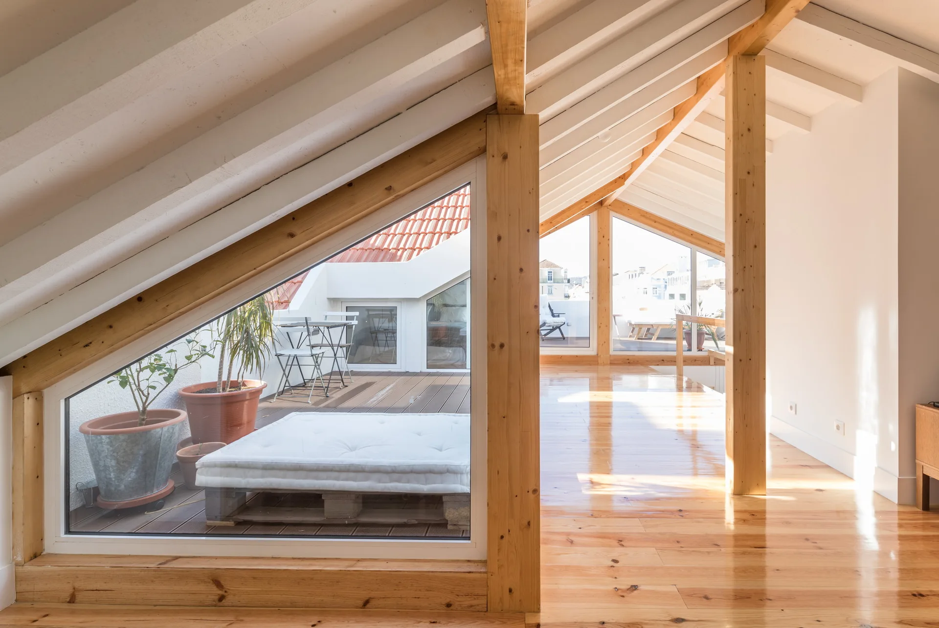 A bright attic room with polished wood floors, exposed structural beams on a sloped ceiling, and large triangular windows overlooking an outdoor terrace.