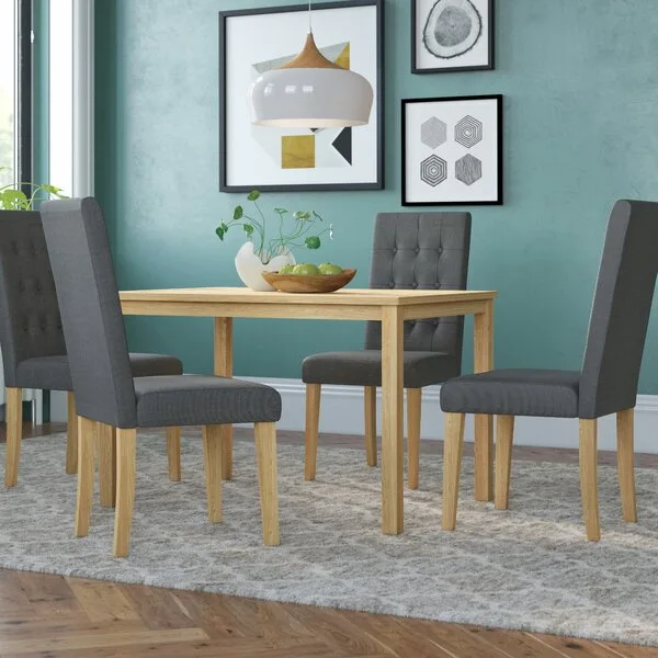 Four grey upholstered tufted dining chairs sit around a light oak table on a patterned rug against a teal wall.