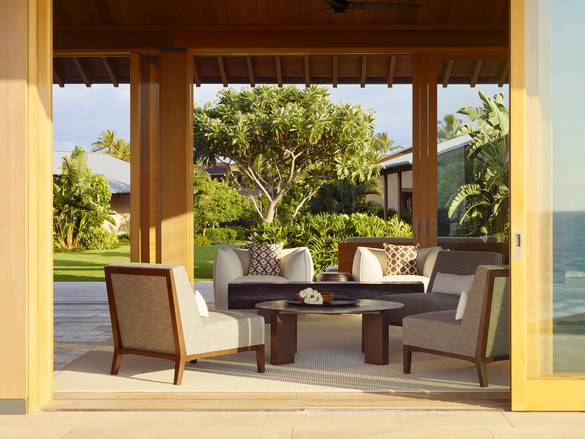 A covered patio features a seating arrangement of armchairs, a wooden coffee table, and an area rug looking out to a lush tropical garden.