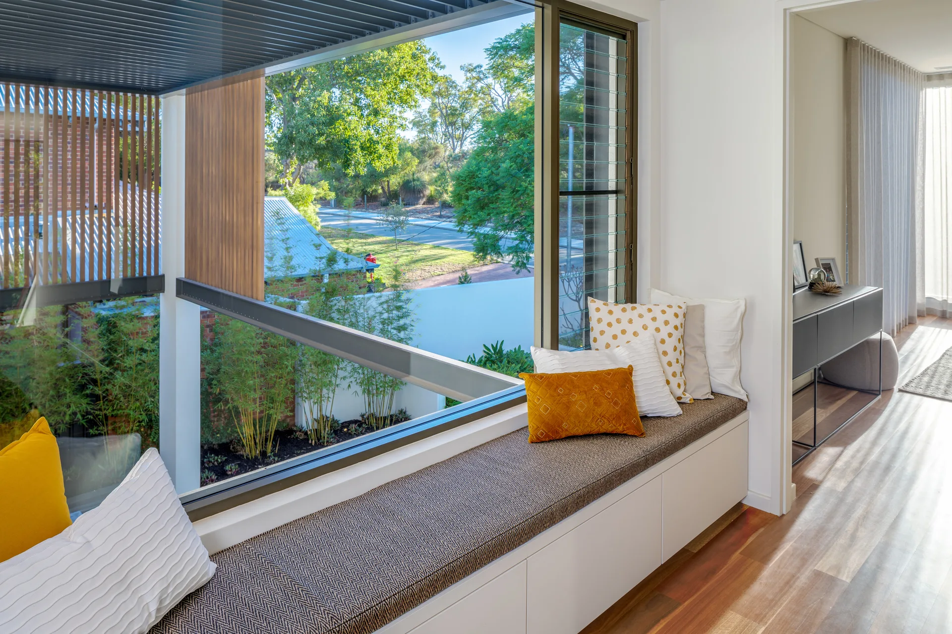 A long built-in window seat with a textured grey cushion and mustard yellow pillows sits beneath a large window overlooking trees.