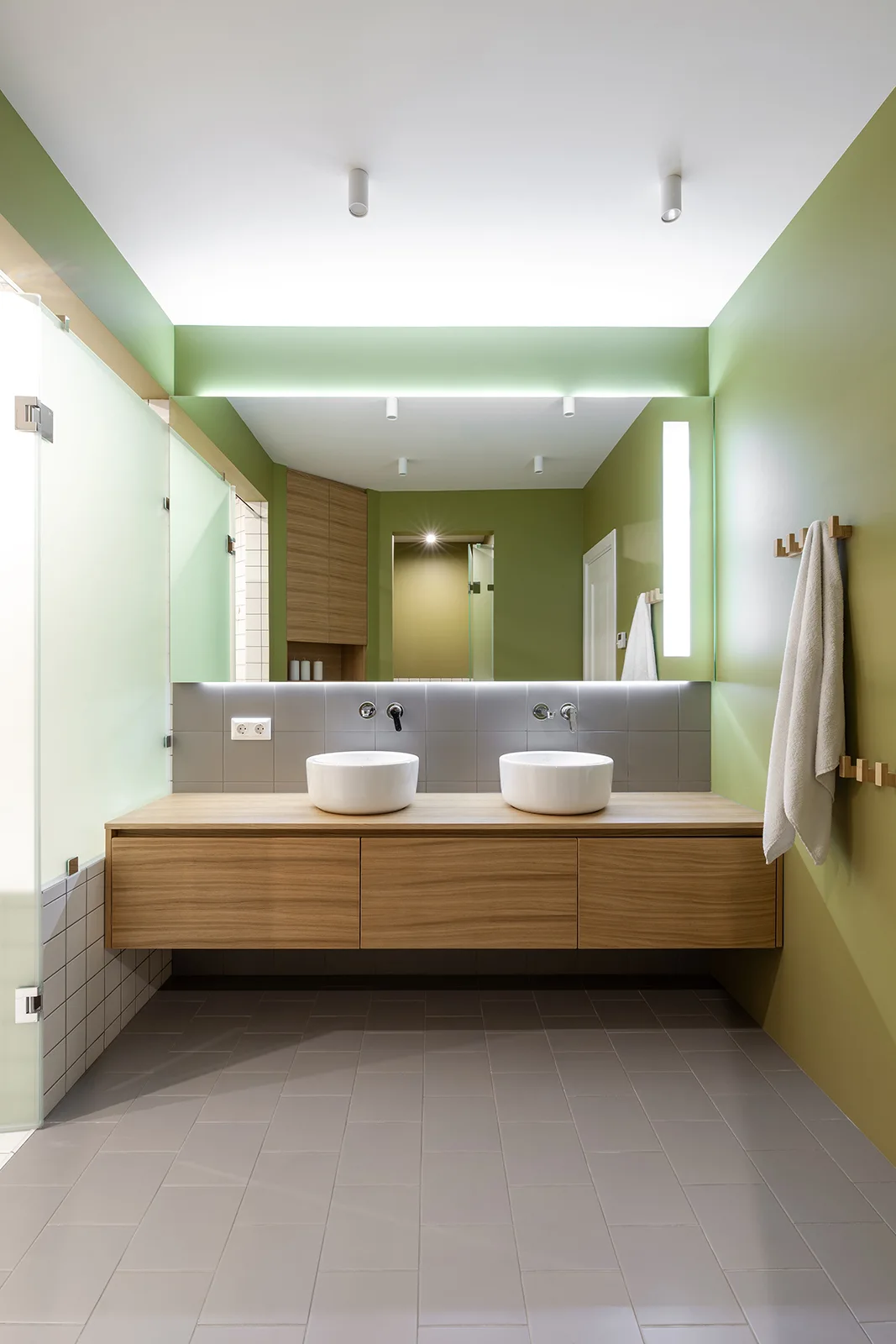 A modern bathroom featuring a floating wood-toned double vanity with two white vessel sinks against sage green walls, a backlit mirror, and grey floor tiles.