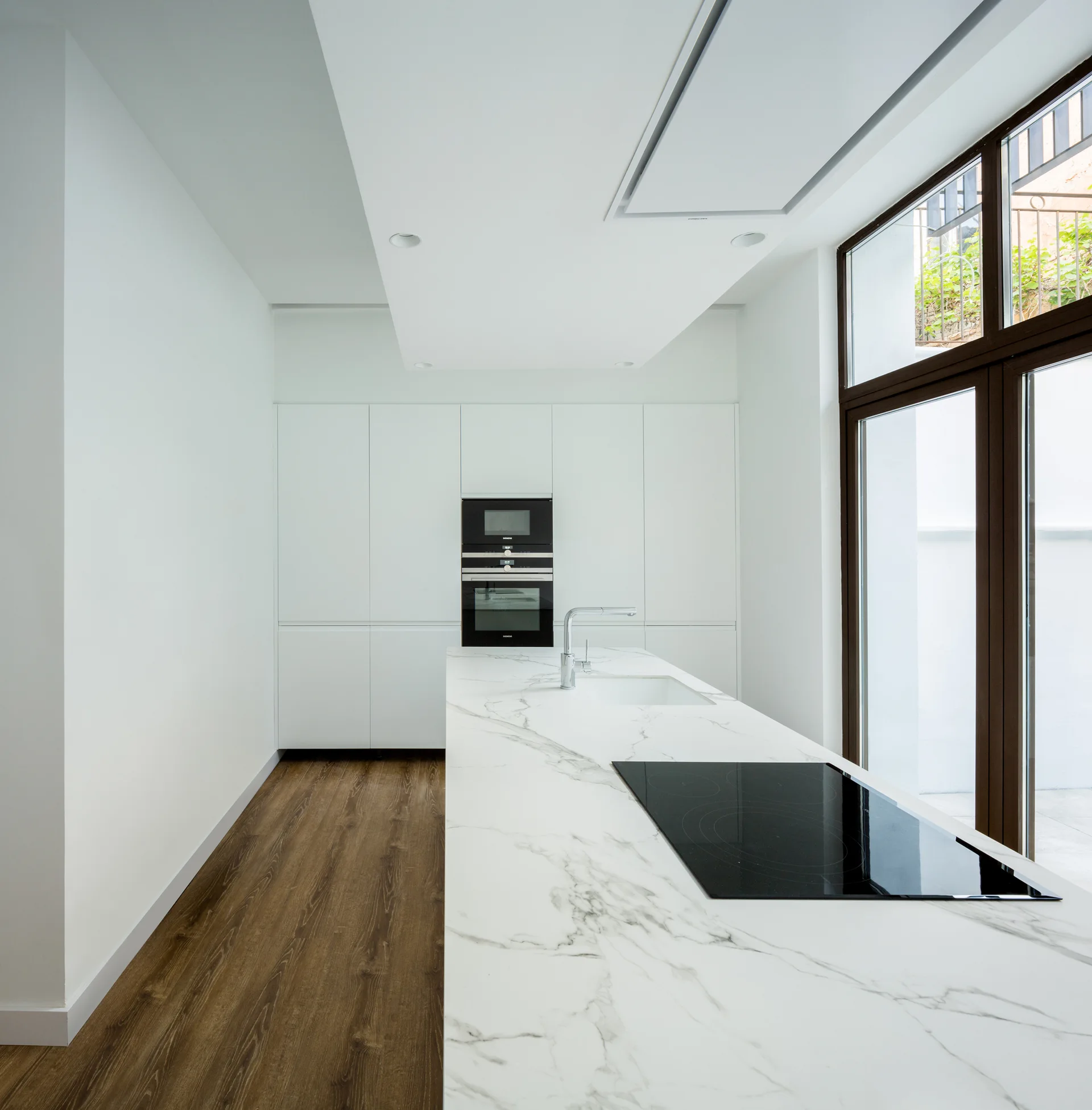 A minimalist white kitchen featuring a long veined marble island, handleless tall cabinets, integrated black appliances, and dark-framed glass doors.