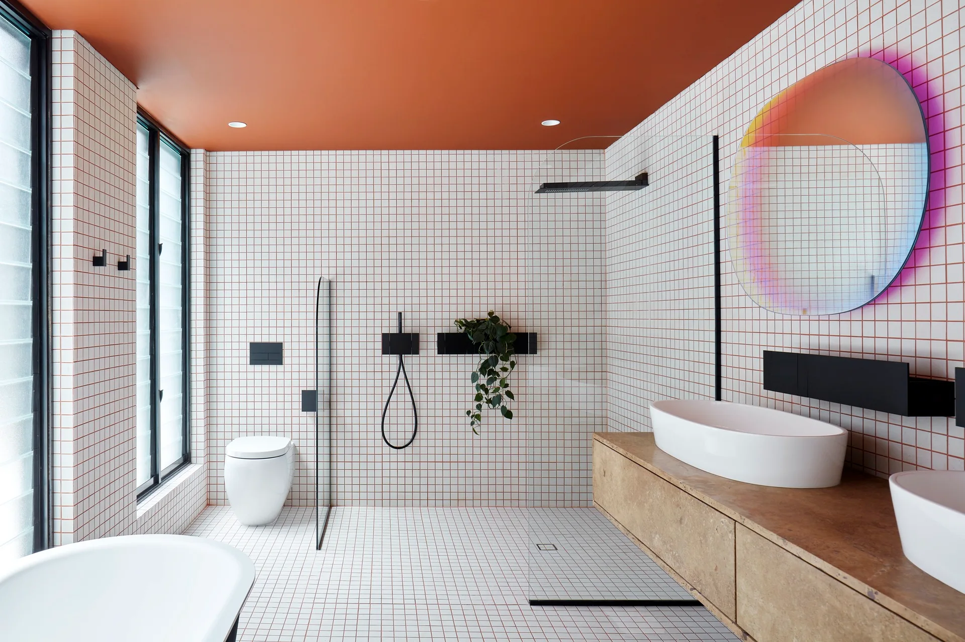 A modern bathroom with white grid tiles on the walls and floor, a terracotta ceiling, a floating wood-look vanity, and a round iridescent mirror.