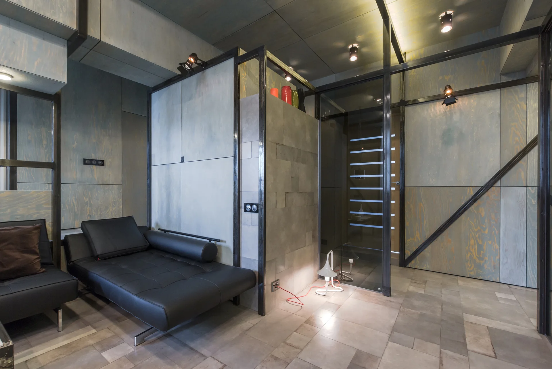 A modern industrial interior featuring blue-stained plywood walls, a black leather daybed, and a glass partition within a black steel frame.